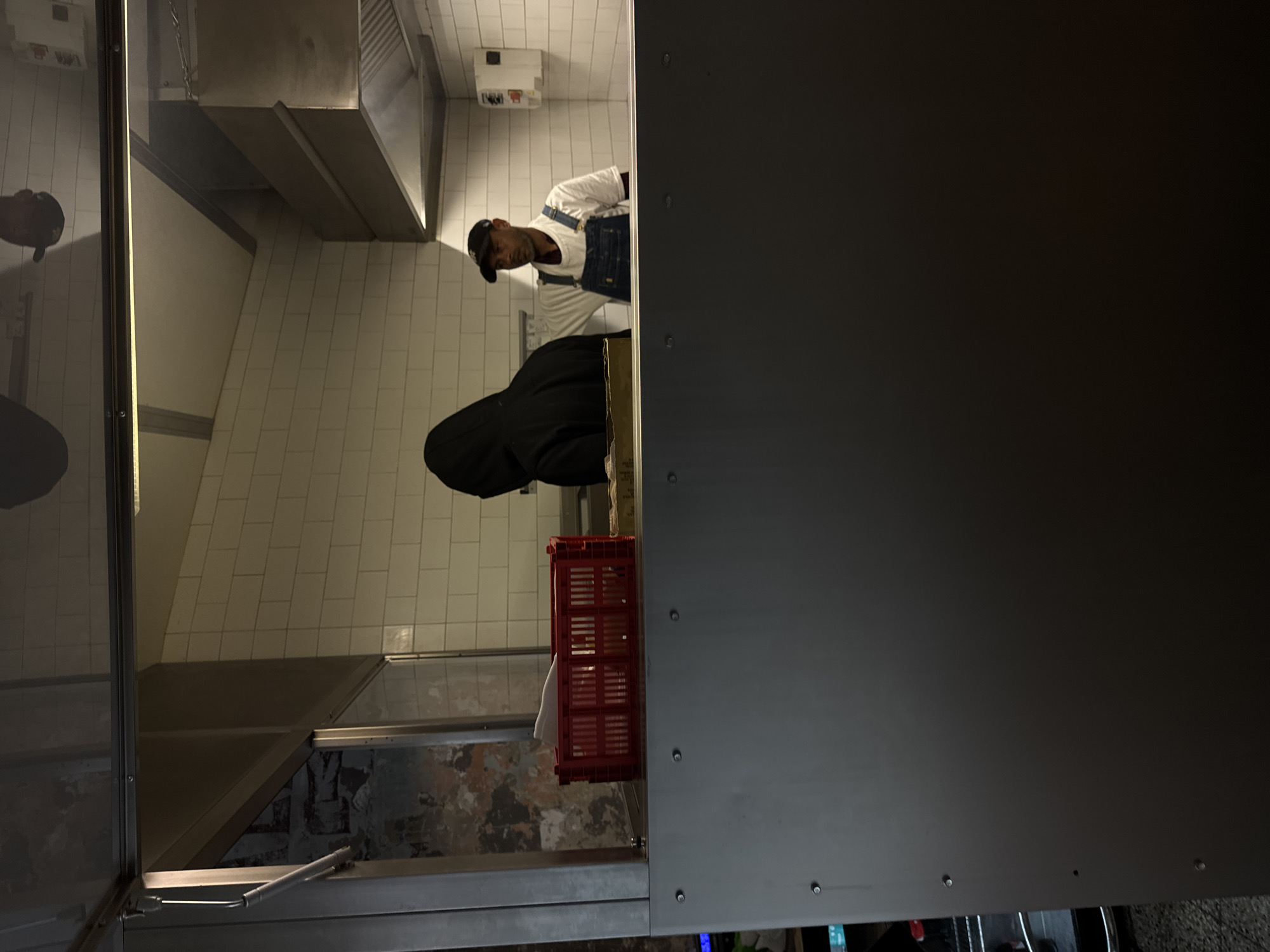The image depicts an indoor scene within a food service establishment in Milan, Italy. The foreground is occupied by a dark gray, riveted metal counter or partition. Above this, a clear glass barrier separates the foreground from a background work area.
To the far left, beyond the glass, a section of an aged wall featuring distressed plaster over exposed brickwork is visible. Immediately behind the glass barrier, on the counter, a red plastic crate contains multiple white folded items.
Two individuals are present in the background work area, which features white rectangular subway tiles on the walls. The person on the left is wearing a dark, hooded garment and is positioned with their back to the viewer, obscuring their face, engaged in an activity at a workstation. To their right stands a male with dark skin, wearing a black baseball cap, a white t-shirt, and blue denim overalls. He has a beard and is looking towards the left, with his left hand on his hip. Above the work area on the right, a stainless steel commercial kitchen hood with visible ventilation grilles is installed, and a white electrical box is mounted on the tiled wall. Overhead lighting is reflected on the glass barrier. The environment indicates a functional kitchen or food preparation space.