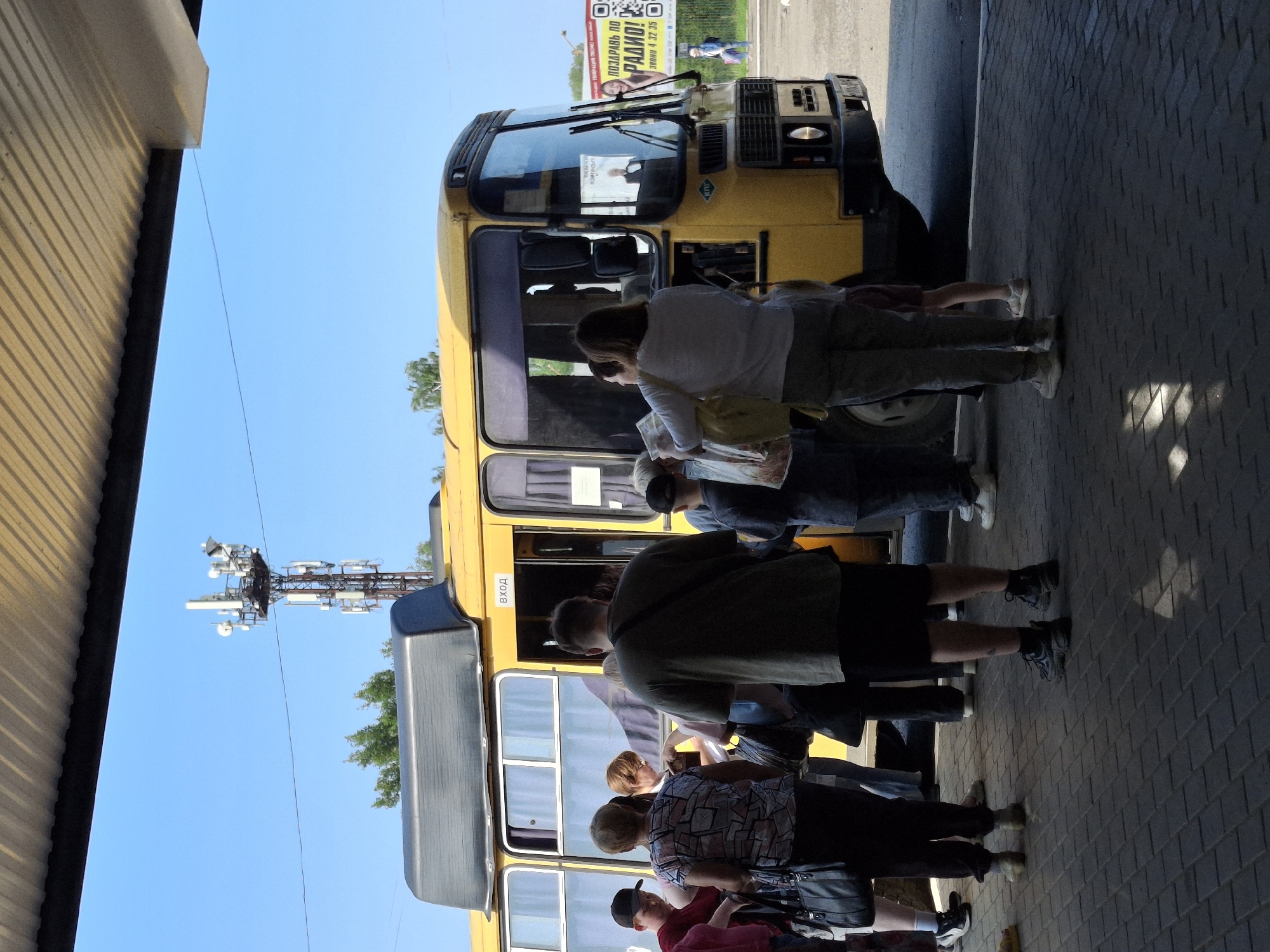 A yellow bus is parked at a bus stop, with several people standing nearby, some appearing to be boarding or disembarking. The bus has its door open, and "ВХОД" (Entrance) is written above it. People are dressed in casual clothing. The environment suggests an outdoor setting under a covered structure, possibly a bus station or shelter, with a clear blue sky visible. A cell tower is prominent in the background. Advertising billboards can be seen in the distance. The ground is paved with interlocking bricks. The scene appears to be in the city of Chaikovsky, Russia, as indicated by the advertising content on a billboard in the background, which features a Russian radio station and promotes an event.
