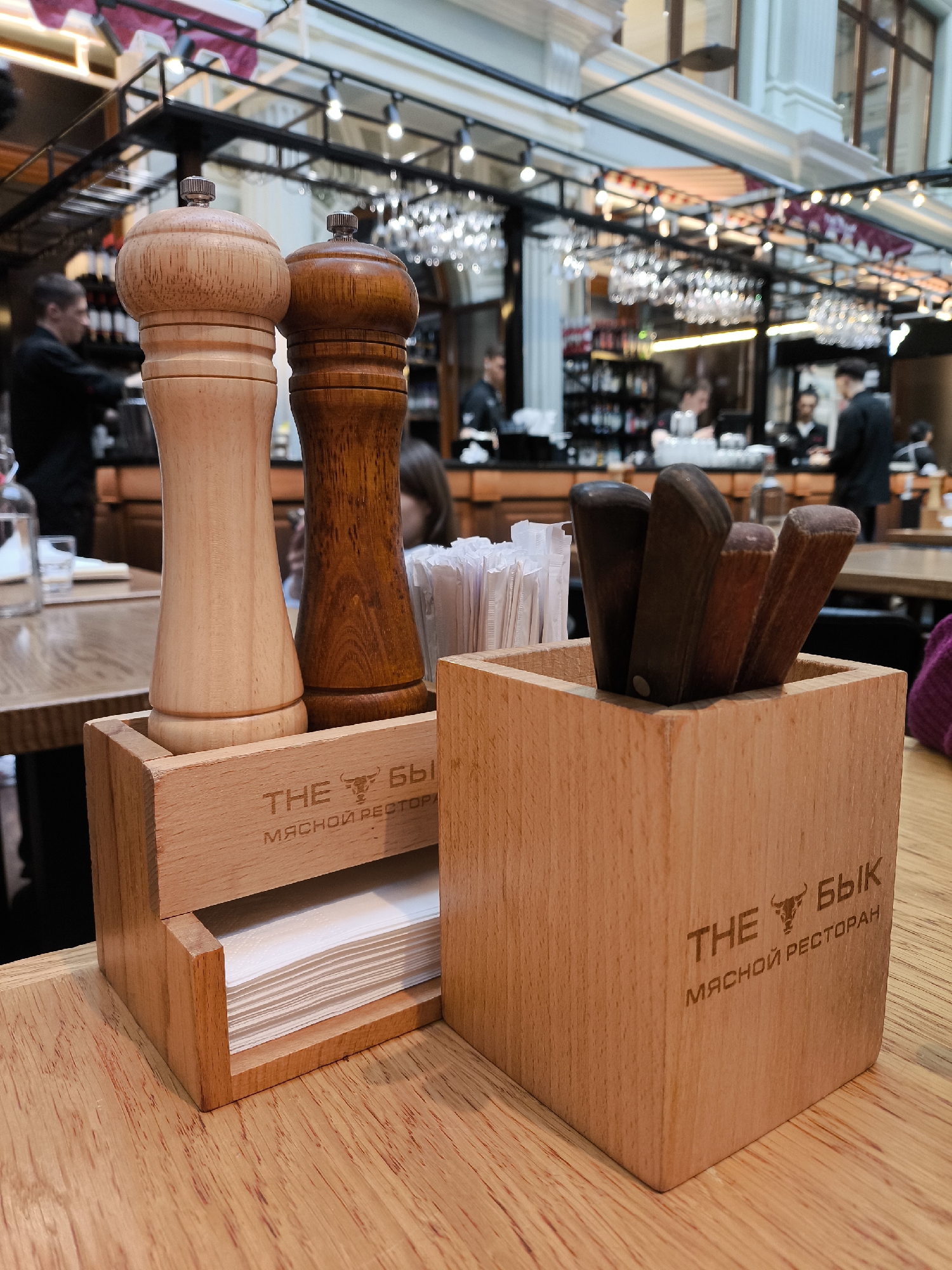 The image depicts a table setting in a restaurant, likely indoors. In the foreground, a wooden container holds a stack of napkins and two wooden salt and pepper shakers. Adjacent to this container is another wooden box containing several steak knives with dark wooden handles. Both containers bear the inscription "THE БыК МЯСНОЙ РЕСТОРАН," which seems to indicate the restaurant's name.
In the background, there is a bar area with staff members and possibly other customers. Above the bar, wine glasses are arranged on racks. The overall lighting suggests a warm, possibly upscale restaurant environment.
The location is stated as being in the city of Unknown, Unknown.