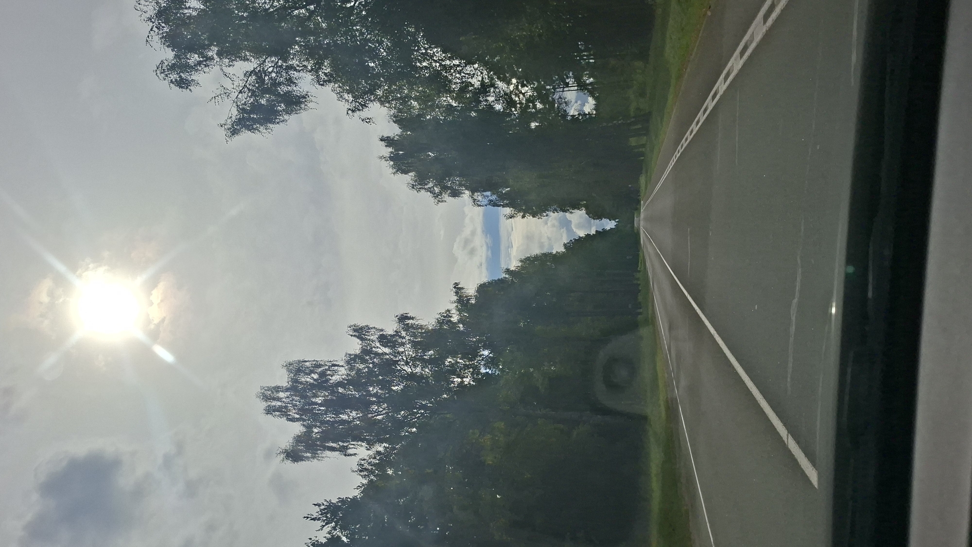 The image captures a straight asphalt road lined with tall, mature trees on either side, creating a tunnel-like effect. The sky above is overcast with bright sunlight bursting through the clouds, creating a lens flare effect. The road has white lane markings and a shoulder. The foreground shows the dashboard of a vehicle, indicating the photo was taken from inside a car. The environment appears to be rural or a scenic route, possibly located within the Мядзельскі сельскі Савет in Belarus, given the presence of birch trees which are common in the region. There are no people or animals visible in the image. The overall impression is of a journey through a wooded landscape on a bright, cloudy day.