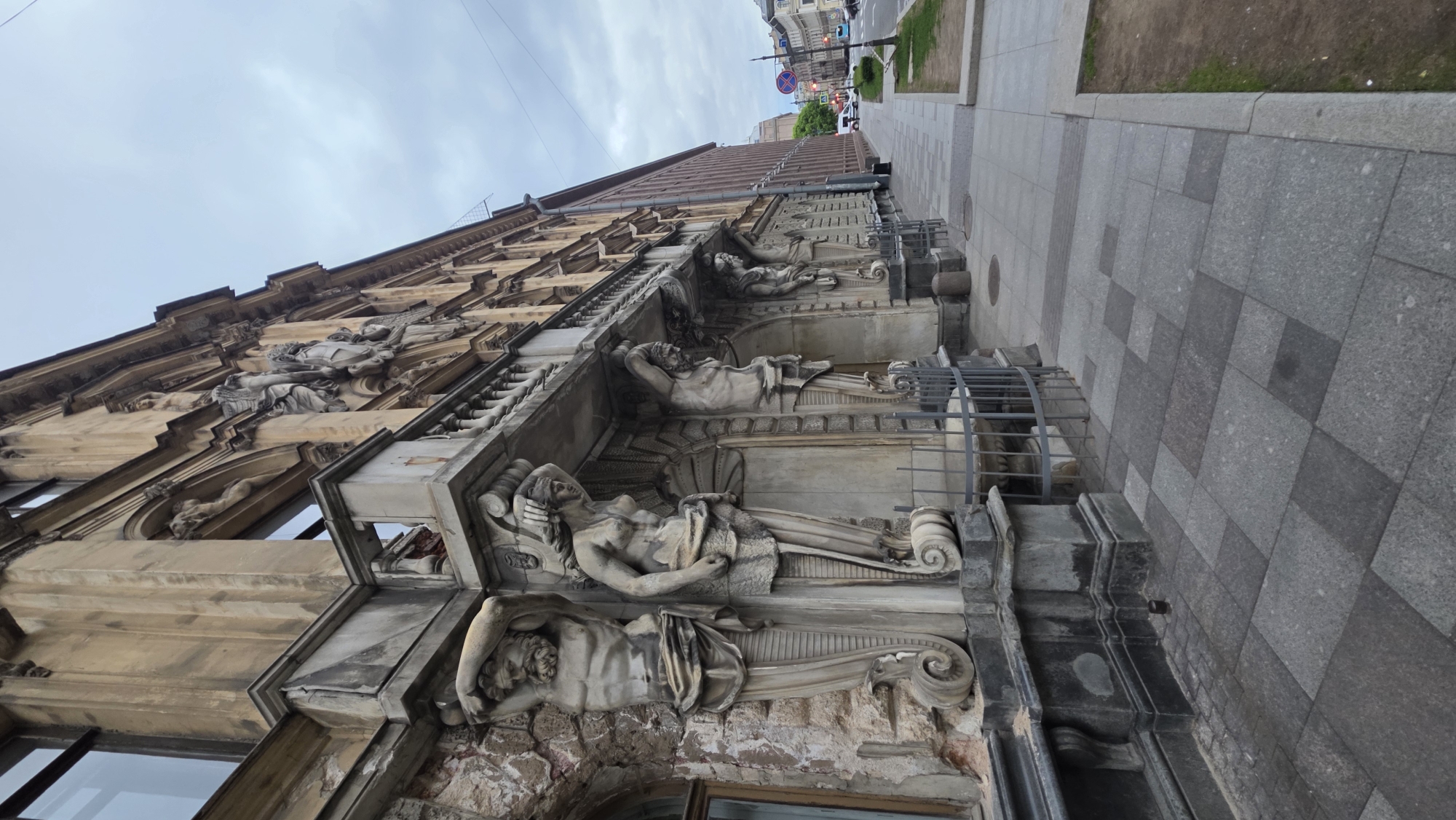 The image shows a detailed view of a building facade in Saint Petersburg, Russia. The building features classical architectural elements, including sculpted figures acting as supporting caryatids or atlantes. The sculptures depict reclining human forms in various poses and attire, appearing to be crafted from stone or a similar material. The facade is adorned with ornate moldings and decorative details. The building appears to be aging, with some signs of wear and disrepair visible. A sidewalk or pavement is visible on the right side of the frame. A glimpse of the street with traffic signs and vehicles can be seen in the distance. The sky is overcast, suggesting diffused lighting conditions.