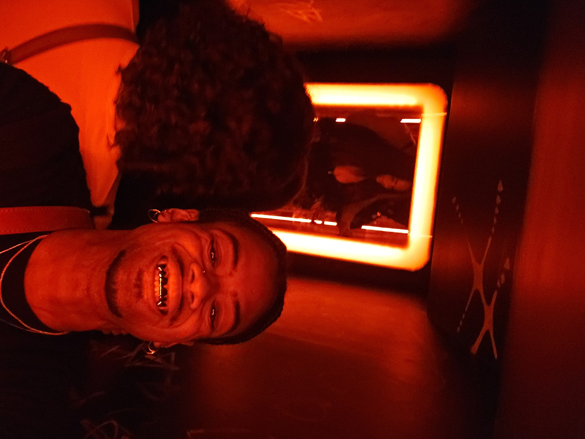 A person is illuminated by red light in what appears to be a narrow space. The individual is wearing a black shirt and has curly hair. The person is grinning and wearing gold grillz. Behind the person is an illuminated rectangular frame. The environment is dark, and a design resembling two crossing strokes or lines is visible on the wall. There are no verifiable indicators in the image to pinpoint the location as Paris, France.