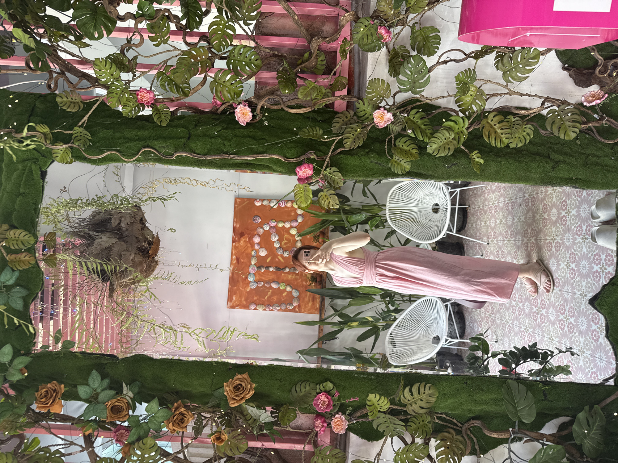 A full body shot of a woman in a pink dress taking a photo of herself in front of a flower wall in Nha Trang ward, Vietnam. She is in a mirrored hallway, and there are two white chairs on either side of her. A piece of art hangs on the wall behind her. The flowers are predominantly pink and tan.