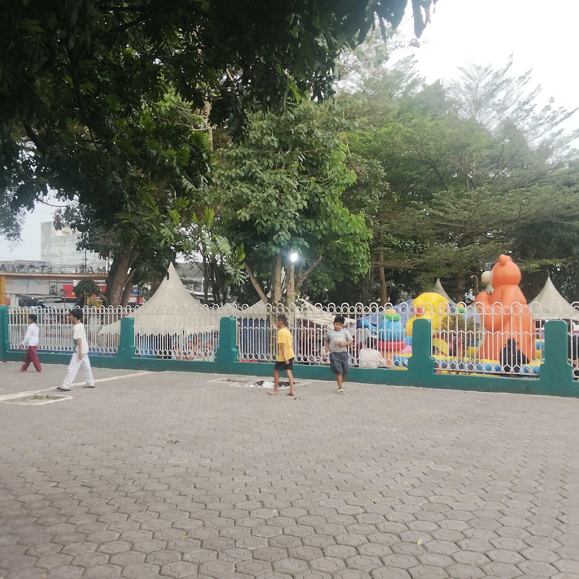 The image depicts an outdoor scene in Cisaat, Indonesia, possibly a park or recreational area. In the foreground, a paved walkway with hexagonal interlocking stones extends into the frame. Several children are present in the mid-ground. Two boys, one in a yellow shirt and black shorts, and another in a grey shirt and dark shorts, are walking on the paved area. To the left, two more boys are seen walking, one in a white shirt and red pants, and the other in a white shirt and white pants. A white metal fence with decorative elements separates the paved area from a colorful inflatable playground in the background. The playground features large, inflatable structures, including what appears to be an orange bear-shaped figure and a yellow dome. Several white tent-like structures are also visible behind the fence. Lush green trees with dense foliage dominate the upper portion of the image, providing shade. In the distance, buildings with white exteriors are visible, suggesting an urban or semi-urban setting. The sky is overcast.