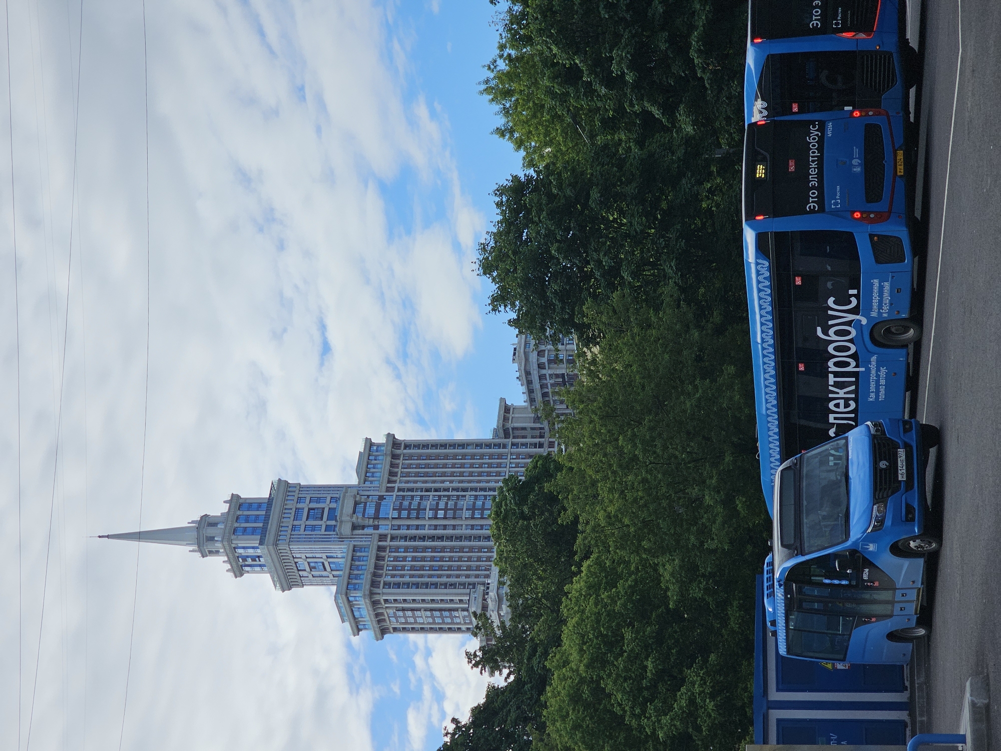 The image captures a scene in Khimki, Russia, featuring a tall building, trees, buses, and overhead wires under a partly cloudy sky. A light-colored building with multiple floors and a pointed spire stands prominently. Green trees are visible in the middle of the frame, positioned between the building and the buses. Several blue "elektrobus" electric buses are parked or moving on a road, with some details and writing visible on their sides. Overhead wires are strung across the top left portion of the image.