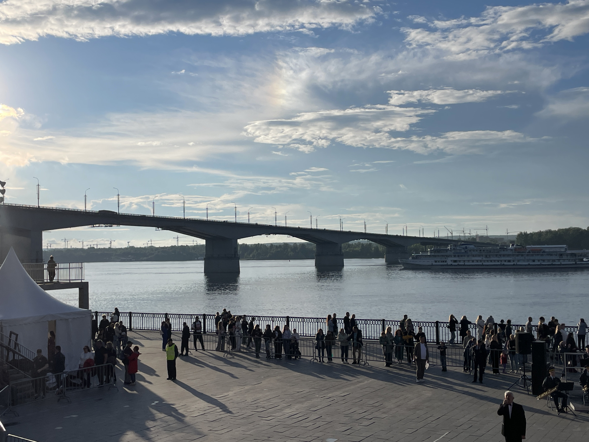 The image shows a wide, paved embankment along a river, likely the Kama River in Perm, Russia. A large concrete bridge spans the river in the background, with a white passenger ship sailing beneath it. The sky is partly cloudy with a rainbow visible. A crowd of people are gathered on the embankment, some standing near a white tent structure, others lined up along a black metal railing overlooking the water. There is a small ensemble of musicians playing in the foreground, with one man in a suit standing apart and appearing to speak into a microphone. The sun is low in the sky, casting long shadows across the ground.