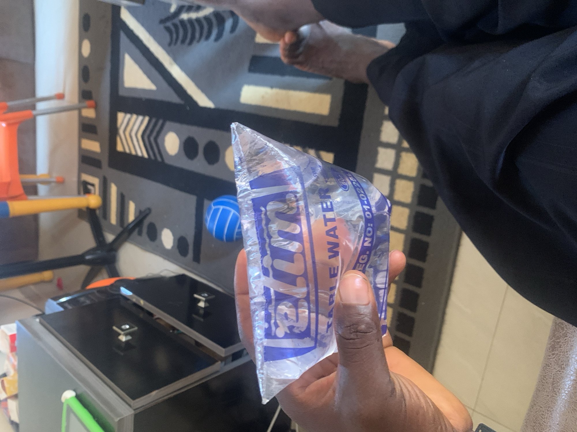 In an indoor setting, likely a room in Gura Top, Nigeria, a person's dark-skinned hand is holding a clear plastic sachet of "TABLE WATER" with blue lettering. The sachet is partially filled with water and is being squeezed. In the background, a patterned rug with geometric designs in black, grey, and white is visible, along with part of a dark cabinet and a glimpse of a bright orange chair with yellow legs. A blue and white ball sits on the rug. The flooring appears to be tiled. The scene is lit by ambient light, creating some shadows. The focus is on the water sachet and the hand holding it.