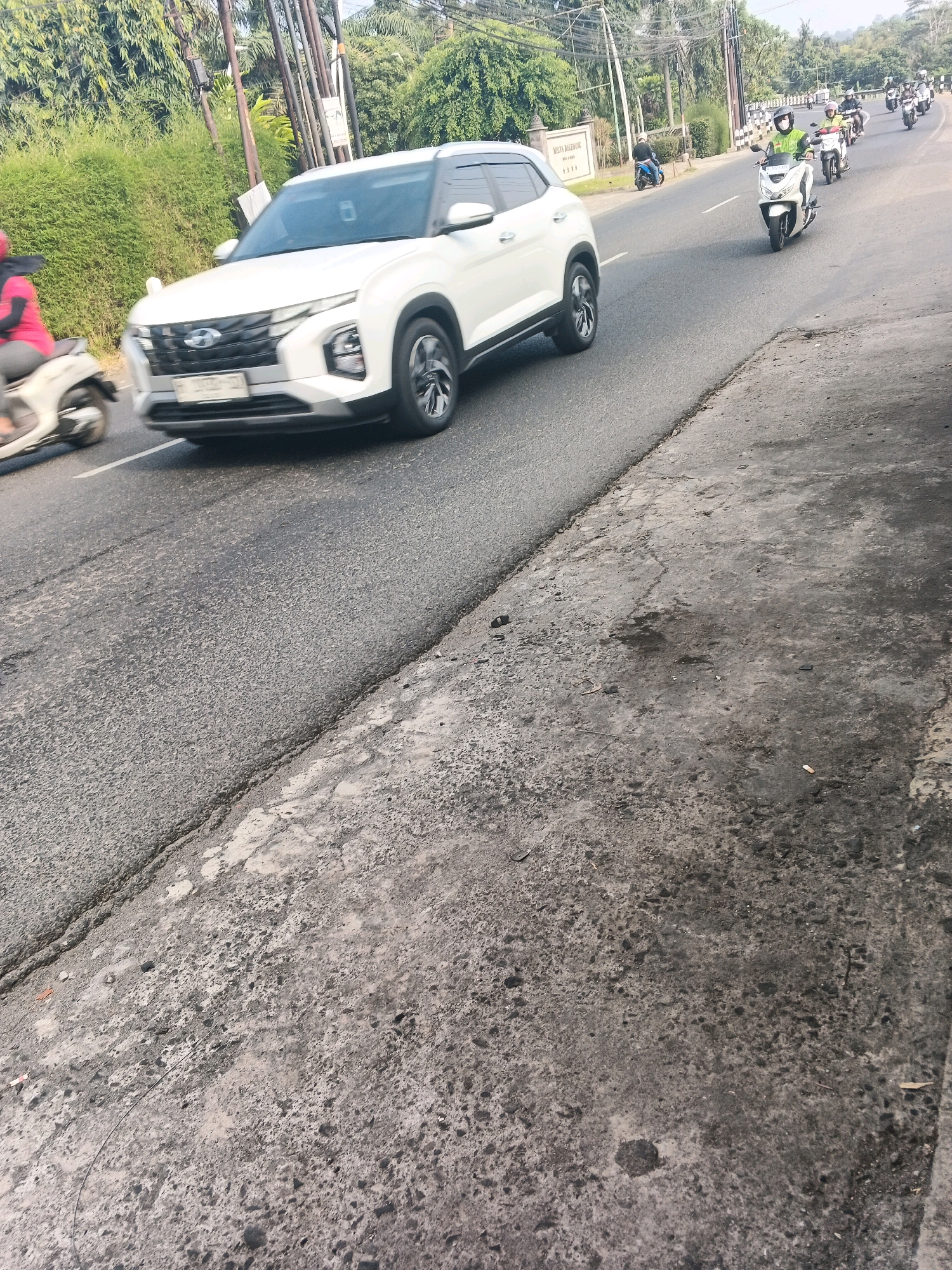 A white Hyundai Creta SUV is traveling on a paved road in Ungaran, Indonesia. The SUV is in the left lane, heading away from the camera. A person wearing a pink shirt and a red headscarf is on a scooter to the left of the SUV, also heading away from the camera. Several other scooters, some with riders wearing helmets, are visible further down the road. The road is bordered by a concrete curb and a grassy area with trees and foliage. There are power lines and poles visible in the background. The road surface is cracked and uneven.