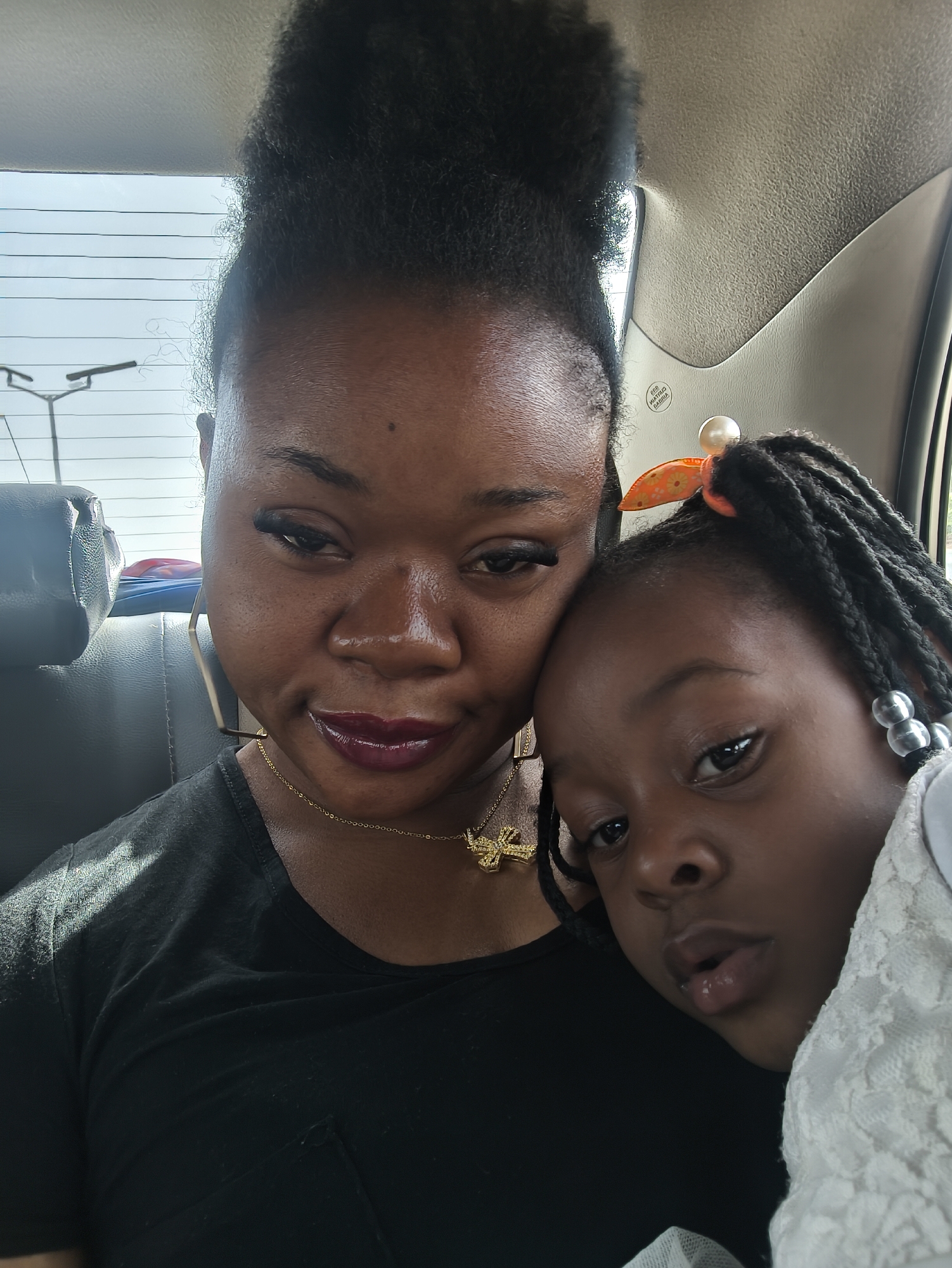 An intimate, bright, and slightly low-angle portrait shows a woman and a young girl sitting together inside a car, likely in Jos, Nigeria, during daylight hours.
The woman, on the left, has dark skin and her dark, curly hair is styled high on her head. She is wearing dark lipstick and appears to have long eyelashes. She wears a simple black t-shirt, a gold chain necklace with a cross pendant, and large, geometric hoop earrings. Her expression is somewhat reflective, with her gaze directed downwards and slightly to the side of the camera.
The young girl, positioned to the right of the woman, is leaning against her, suggesting a close relationship, possibly mother and daughter. She has dark skin and her dark hair is styled in braids adorned with clear or white beads. An orange bow with a pearl-like embellishment is clipped into her hair. She is wearing a white garment, possibly with a lace texture, and looks directly at the camera with a neutral expression.
The setting is the interior of a car, with a dark headrest visible behind the woman and light-colored ceiling lining. Bright sunlight streams in from the windows, illuminating the subjects and the car's interior. Through the car's rear window, a light blue sky is visible, along with distant utility poles or wires. On the car's ceiling lining, faint, reversed text from a label, possibly a warning or instruction, is barely discernible.
