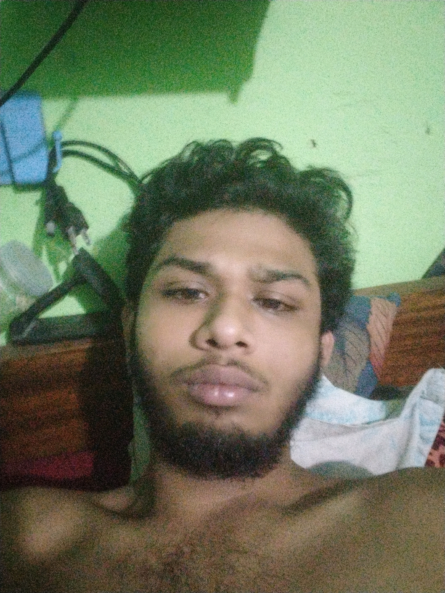 This is a close-up, low-resolution selfie of a young man with dark, curly hair and a beard. He is shirtless and lying down, possibly in bed, as evidenced by a pillow with a patterned case visible behind him. The background wall is painted a bright, uneven green. To the left of the frame, a dark cable and a blue object are visible against the green wall. The lighting is dim, and the image is grainy, suggesting it was taken in low-light conditions, possibly at night. The man is looking directly at the camera. The overall impression is of an informal, personal photograph taken indoors in what appears to be a residential setting. There is no clear indication of the specific location within Dhaka, Bangladesh, beyond it being an indoor urban environment.