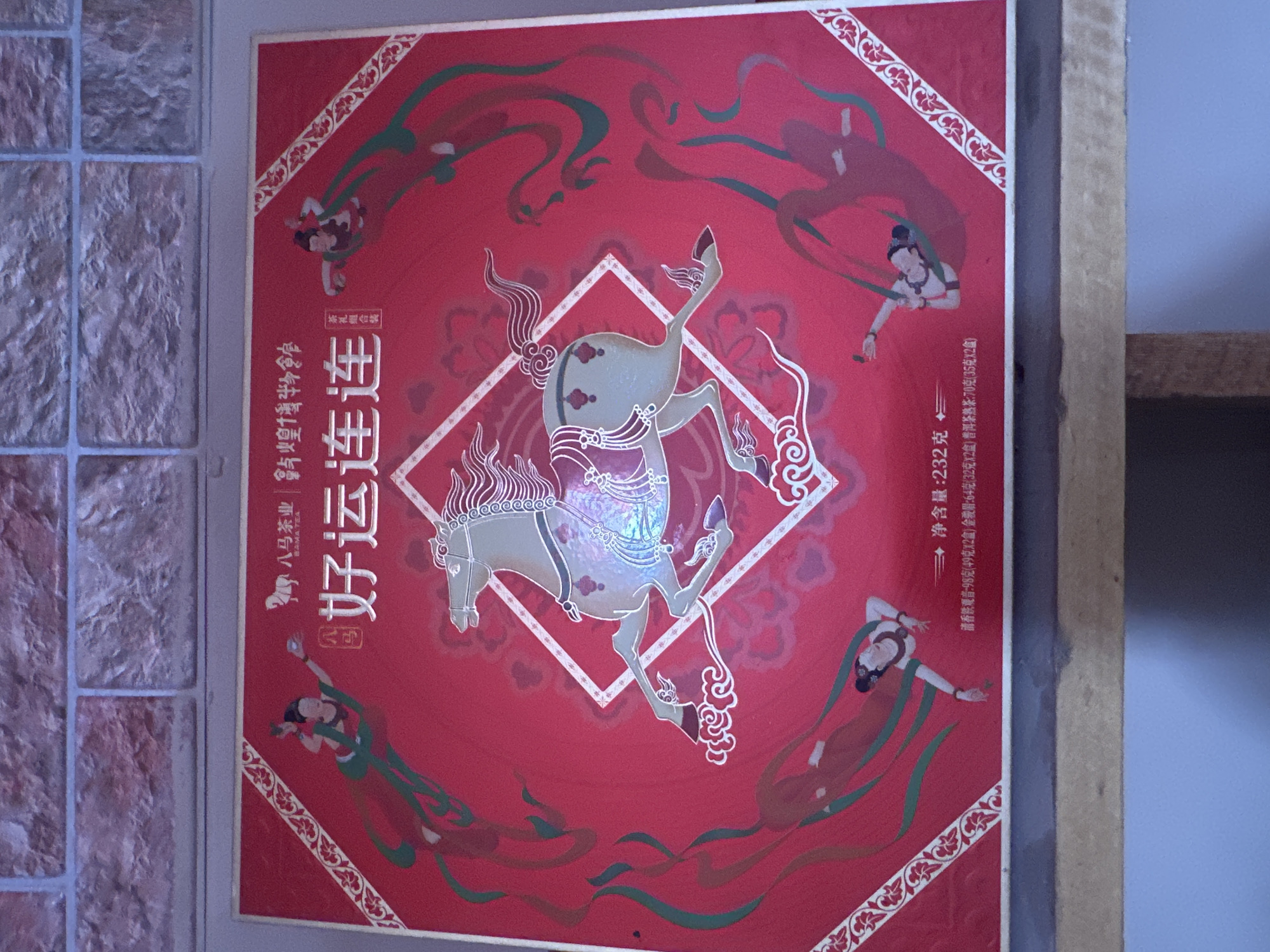 This image shows a close-up of a red rectangular box, likely a gift box for tea, with a festive design. The design features a central image of a grey horse with intricate detailing, possibly representing good luck or prosperity, as suggested by the prominent Chinese characters "好运连连" (hǎo yùn lián lián), meaning "continuous good luck." Around the horse, there are depictions of figures in flowing robes, reminiscent of traditional Chinese celestial beings or dancers, rendered in shades of red, green, and gold. The box is adorned with ornamental patterns along its edges and corners. Text on the box, written in Chinese, indicates the net weight and possibly the contents, suggesting it contains different types of tea. The background is a textured wall made of brick tiles in shades of pink and brown. The box is placed on a wooden surface, suggesting it is on display or being stored. The image's lighting is neutral. The location of this image is not specified within the image itself, therefore it cannot be definitively stated to be in Tashkent, Uzbekistan.