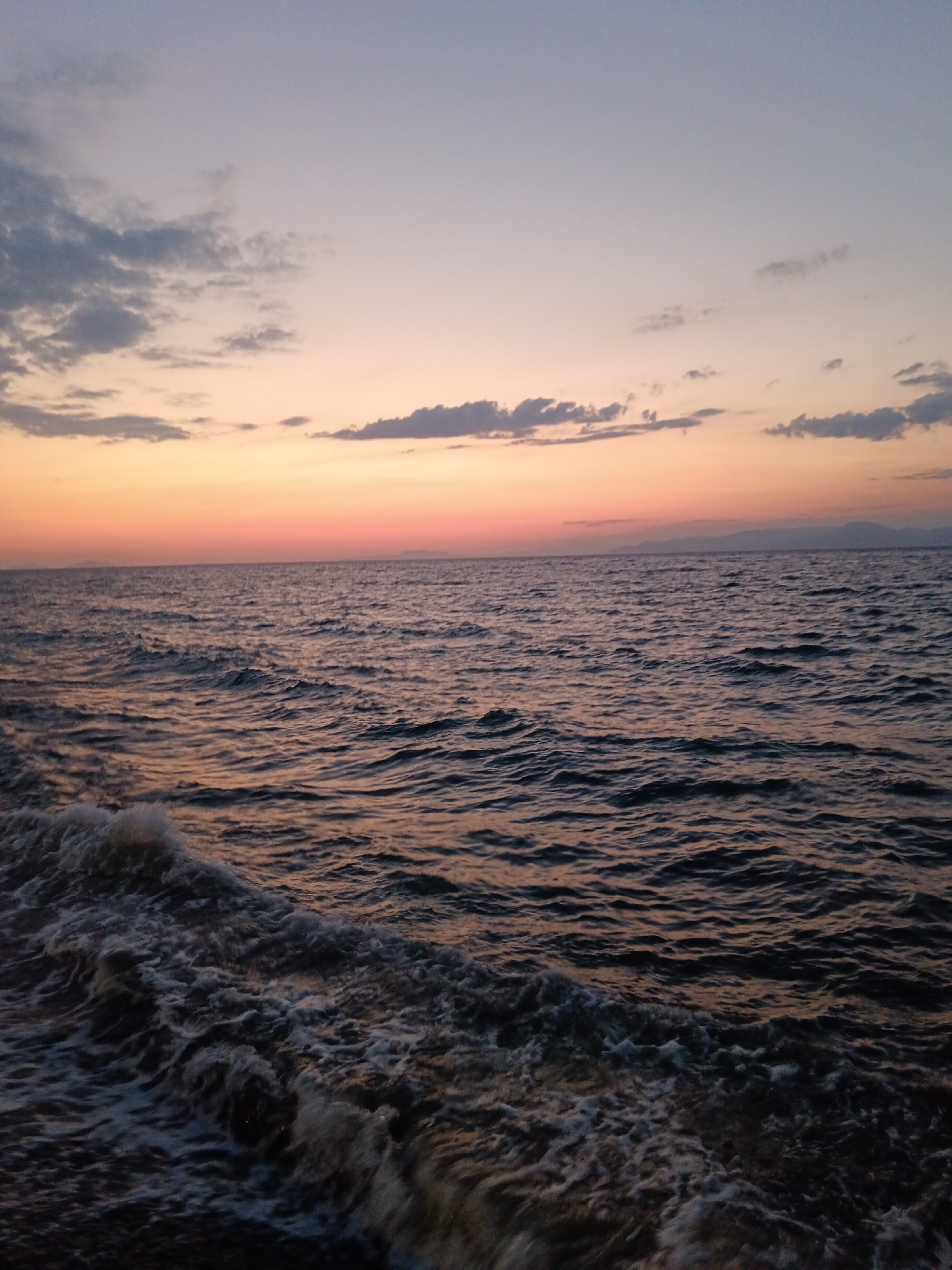 The image depicts a body of water, likely the sea, with waves crashing in the foreground. The water is dark and choppy, reflecting the colors of the sky. The sky above is a gradient of colors, transitioning from a pale blue at the top to hues of orange and pink near the horizon, indicating either sunrise or sunset. Scattered clouds are visible in the sky, some appearing darker and more defined than others. In the distance, a faint landmass or hills can be seen on the horizon. The scene appears to be natural and serene.
The location is the Municipal Unit of Vrachneika, Greece. This is a coastal area known for its natural beauty and proximity to the sea. The specific scene captured in the image is likely a view from the coastline or a boat at sea, showcasing the natural environment of Vrachneika during the twilight hours. There are no people or man-made structures prominently visible in the image, emphasizing the natural elements of the seascape.