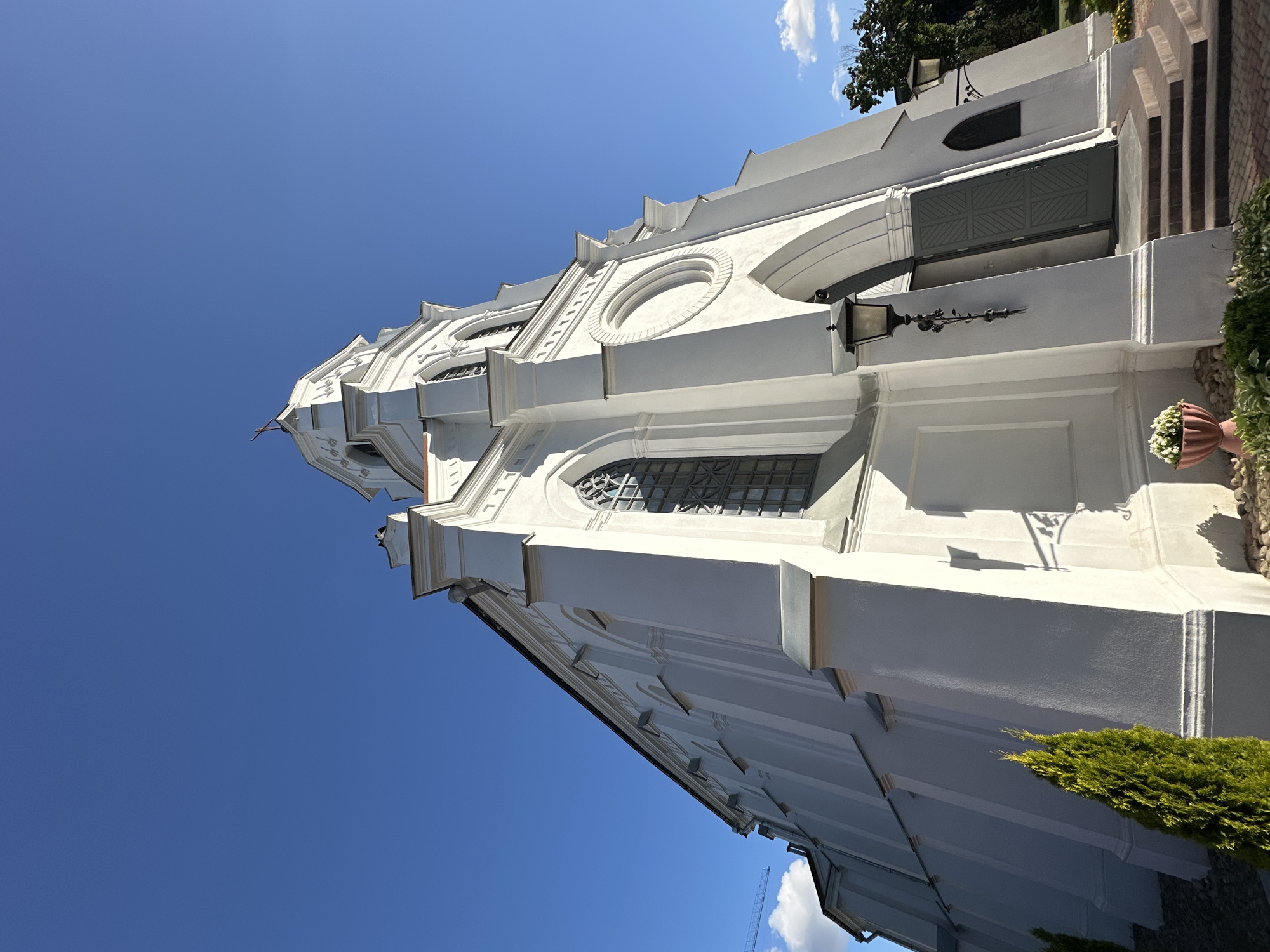 This is a low-angle exterior shot of the Church of Saints Simon and Helena in Minsk, Belarus, under a clear blue sky. The imposing white facade of the church dominates the frame, with its pointed spires, arched windows, and decorative architectural elements. The building exhibits Neo-Gothic and Romanesque influences, characterized by its clean lines and stark white coloration. The entrance features a pair of gray double doors, slightly ajar, revealing darkness within. A black wrought-iron lantern hangs to the left of the entrance. Steps lead up to the doors, and the foreground is landscaped with gravel, a small evergreen tree on the left, and a pink planter containing white flowers on the right. The overall impression is one of solemn grandeur.