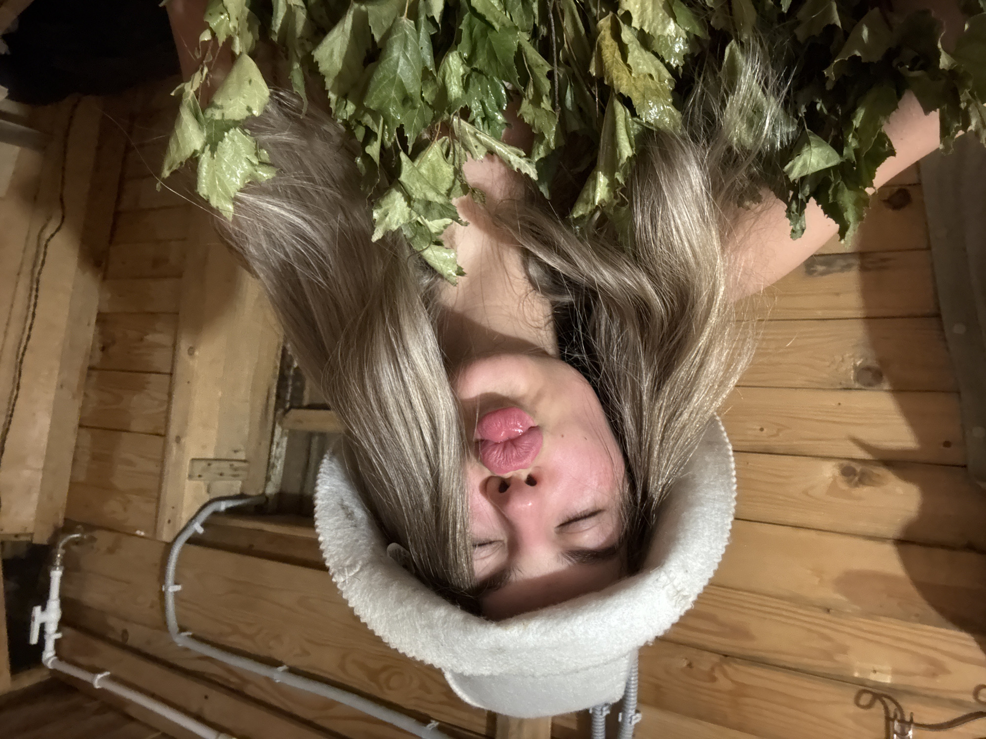 The image shows a woman in a sauna. She is wearing a white sauna hat and is surrounded by birch branches. The walls of the sauna are made of wood. In the lower left there is a set of pipes with a valve.
The city of Chaikovsky, Russia, is not discernible from the image.