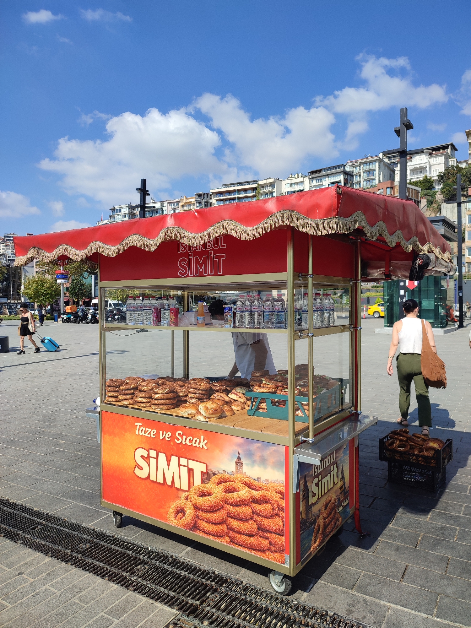 In the city of Beyoğlu, Turkey, a bright red simit cart stands prominently on a paved public square under a clear blue sky dotted with white clouds. The cart's canopy is adorned with gold fringe, and the front displays vibrant advertisements featuring stacks of simit bread and text that reads "Taze ve Sıcak Simit" (Fresh and Hot Simit) and "Istanbul Simit." Inside the glass-enclosed cart, rows of simit loaves are neatly arranged on shelves, alongside bottled water.
To the left of the cart, a woman wearing a black dress and sunglasses walks away from the camera, pulling a bright blue suitcase. In the background, several motorcycles are parked, and buildings rise up a hillside.
To the right of the cart, a woman wearing a white tank top and green pants walks away from the camera, carrying a brown fringed bag over her shoulder. A black crate filled with simit bread sits on the ground to her right, casting a distinct shadow. The square is paved with large, grey stone tiles, and a metal grate runs along the foreground, also casting shadows. The overall scene depicts a typical street scene in a bustling Turkish city, highlighting a popular local food item.