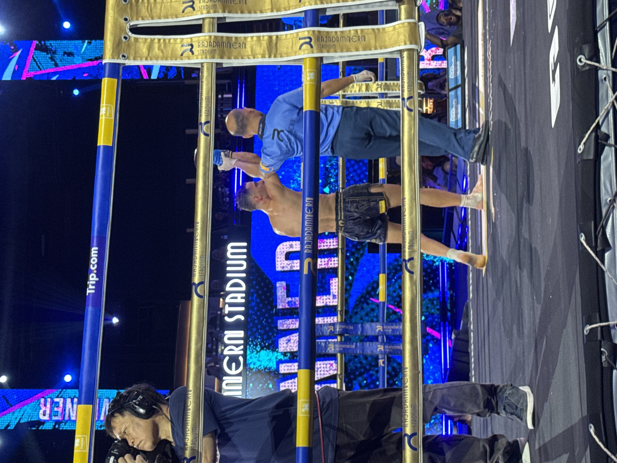 The image captures a scene at Rajadamnern Stadium in Bangkok, Thailand. A shirtless fighter is being checked by a referee. A cameraman stands at the base of the boxing ring. Banners and ring posts with "Rajadamnern Stadium" are visible, and the "Trip.com" advertisement appears on a blue post. The background displays a colorful LED screen. The ring is black.