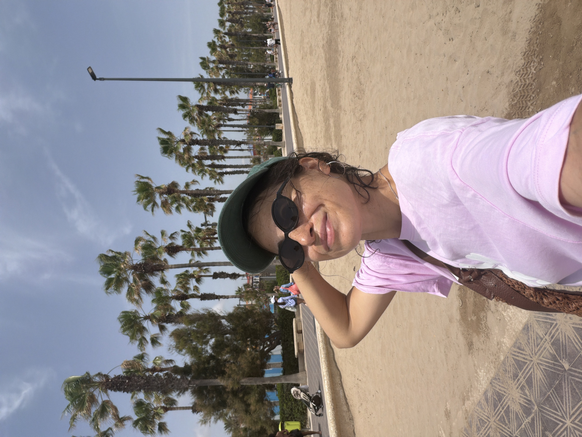 A woman with dark hair, wearing sunglasses, a green baseball cap, and a pink t-shirt, is taking a selfie on a sandy beach. She is smiling and has her hand on the back of her head. In the background, there are rows of palm trees, a street, and several people walking and riding bicycles. The sky is clear and blue with a few wispy clouds. The beach has a paved walkway with a geometric pattern and a sandy area.