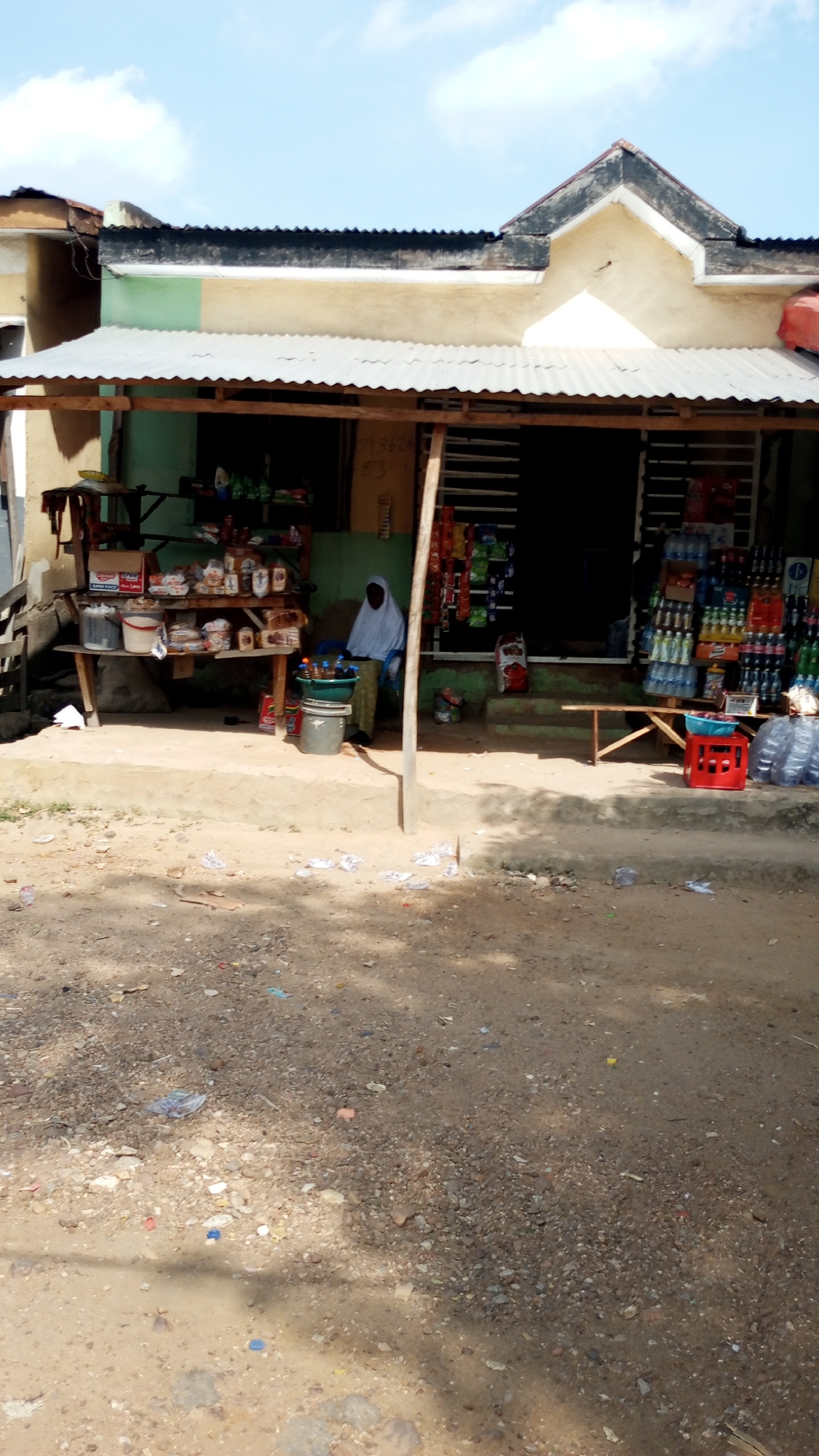 The image depicts a small retail establishment in Adankolo, Nigeria. The shop is housed in a single-story building with a weathered beige exterior, green trim, and a corrugated metal roof extending to form a covered storefront. A woman wearing a white hijab is seated inside the shop.
The shop's display consists of wooden shelves stocked with packaged goods, food items, and bottled beverages. Assorted drinks are displayed on a wooden bench and red plastic crate. The ground in front of the shop is dirt-covered and littered with scattered trash.