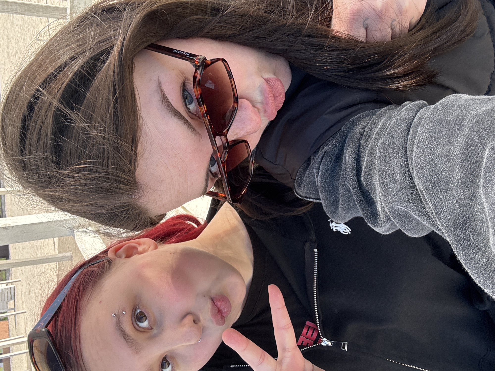 Two individuals pose closely for a selfie. The individual at the top has brown hair and is wearing sunglasses. They are making a face by sticking out their tongue. The second person has red hair and is making a peace sign with their hand. A gray sweatshirt is visible. The location appears to be an outdoor setting, but the city of Chornomorskoe, Ukraine, cannot be confirmed based on this image.
