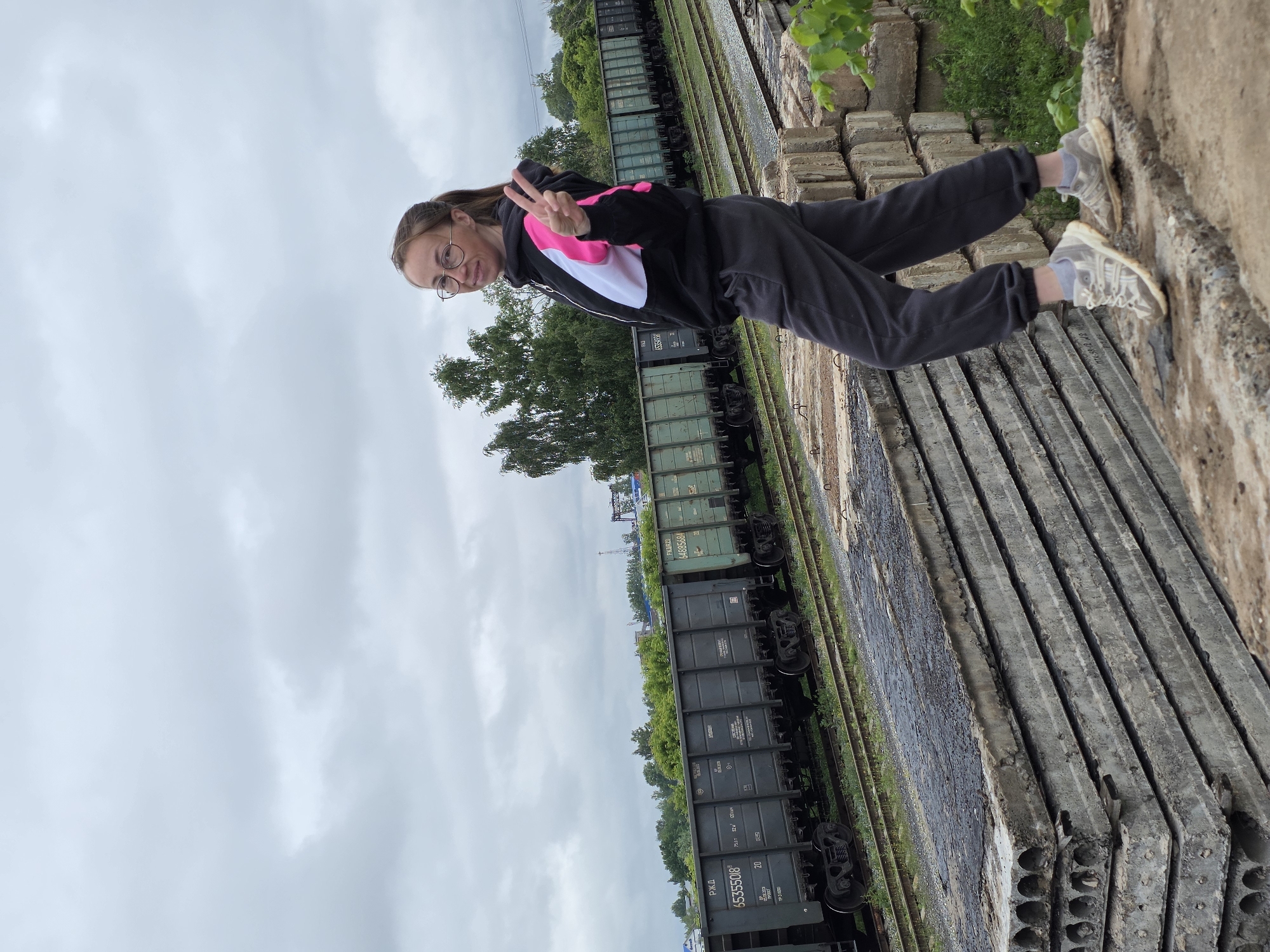 An outdoor scene in Chaikovsky, Russia, depicts a young adult female standing on a stack of weathered concrete slabs under an overcast sky. The female has brown hair tied in a ponytail, wears round-framed glasses, a black hoodie with pink and white sections, dark grey sweatpants, and athletic shoes. She is smiling and making a peace sign with her right hand. Behind her, multiple train tracks feature a line of dark green Russian Railways (РЖД) freight cars, including identifiable numbers such as 65355018 and 64885684. Dense green foliage and trees form the background, with distant industrial structures faintly visible under the uniformly grey, cloudy sky. The concrete slabs in the foreground are stacked, displaying circular voids and dark, possibly asphaltic, residue on their exposed surfaces.