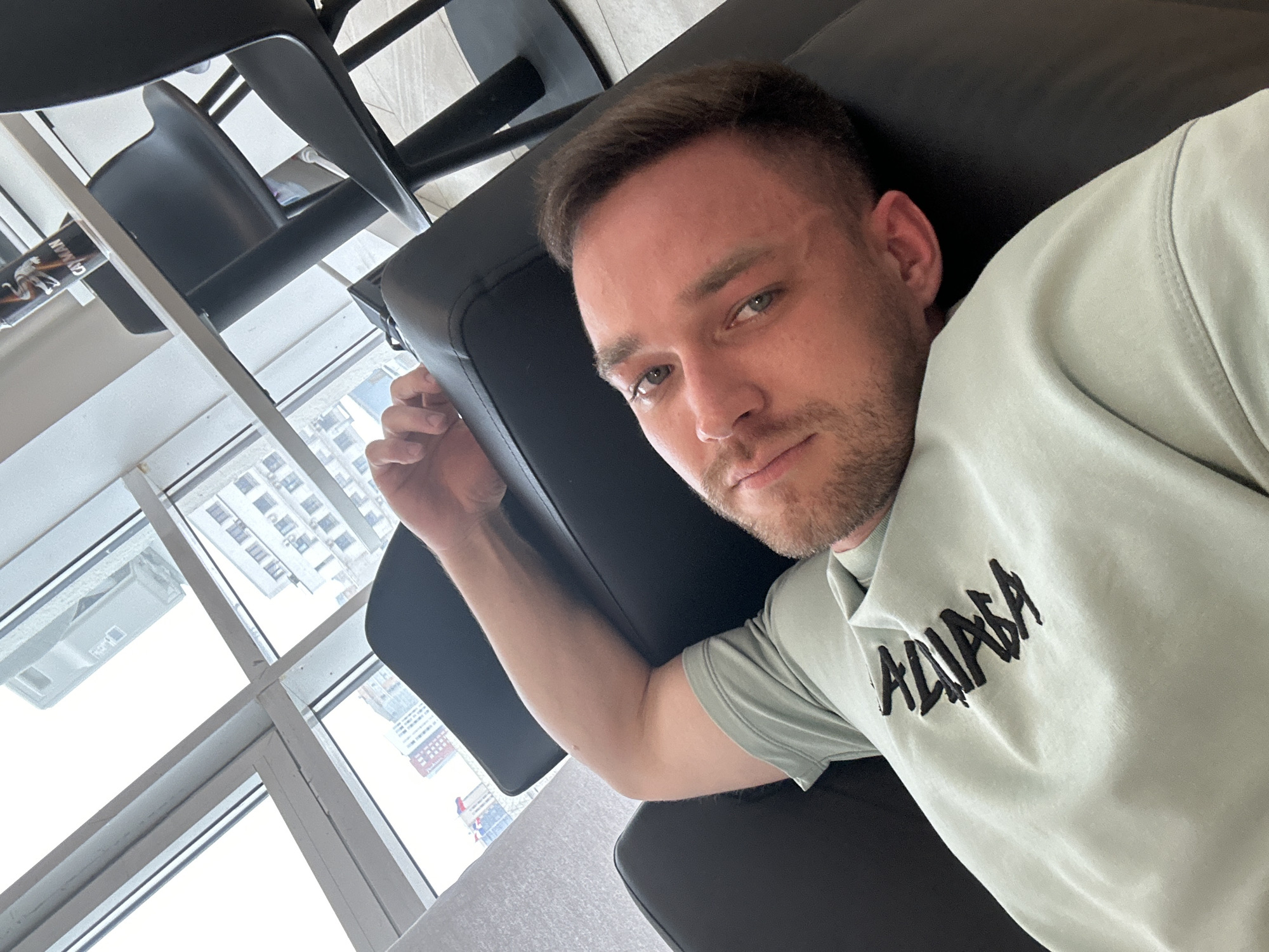 A man with short brown hair and a light beard is reclining in a black leather chair. He is wearing a light green shirt with dark lettering on the right side of the frame. To the left, behind the man and chair, is a large window with a view of city buildings. Based on the architecture, this is most likely in Perm, Russia. The window frame is white. There are two additional black chairs visible above the man. The scene is well-lit.