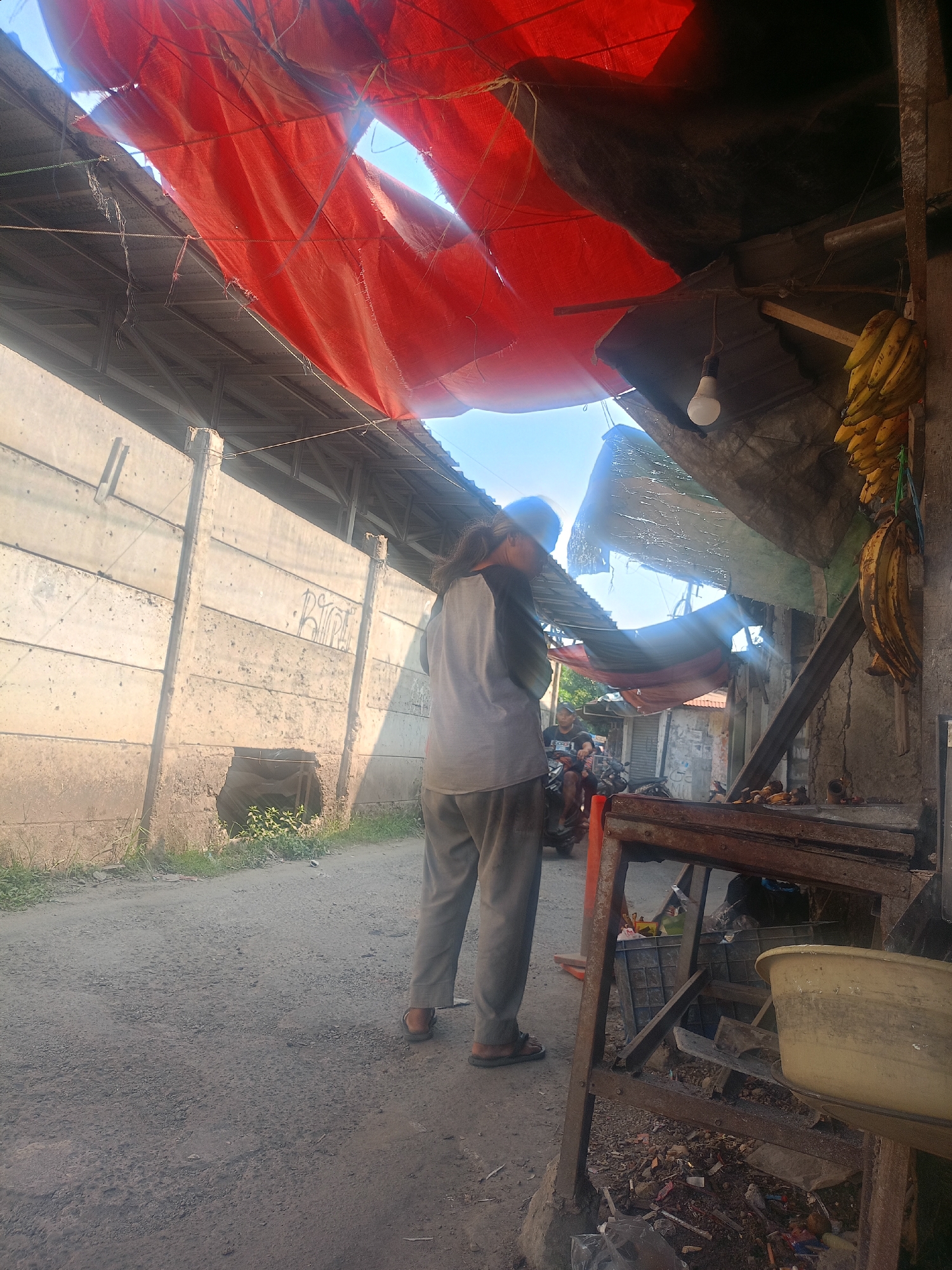 The image depicts a street scene in Bojong Gede, Indonesia, under a makeshift canopy of red and black tarpaulin. A person wearing gray pants and a light-colored shirt stands facing away from the camera, looking towards a motorcycle in the middle ground. Behind them, a concrete wall runs along the left side of the scene, with some graffiti visible. To the right, a rustic stall is set up with a metal frame, displaying a bunch of yellow bananas hanging from a string and various items on a shelf. The ground is a dirt road covered in gravel and debris. The overall impression is one of a humble, possibly outdoor market or roadside vendor setup in a somewhat weathered urban environment.