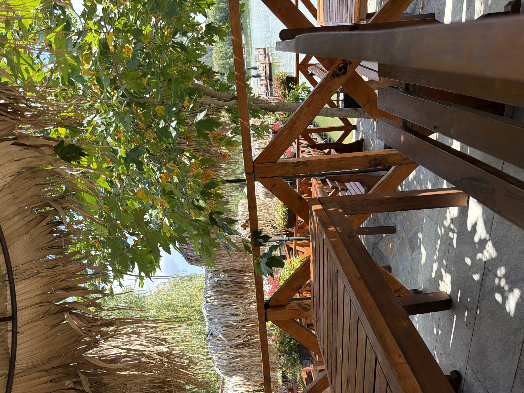 The image depicts an outdoor seating area at a waterfront restaurant in Gorna Malina, Bulgaria. The foreground is dominated by wooden tables and benches, arranged on a paved patio. A wooden railing, constructed from crisscrossed beams, separates the seating area from the view of the water. Overhead, a thatched roof provides shade, composed of dried palm leaves or similar vegetation.
In the midground, a tree with green leaves and some yellowing foliage stands behind the railing. Beyond the tree, a calm body of water is visible, with a small floating platform in the distance that appears to be another seating area or dock, equipped with tables and umbrellas. The background shows a treeline and a hazy sky. The overall impression is of a tranquil, natural setting, likely designed for outdoor dining and relaxation. There are no people visible in the image. The lighting suggests a sunny day, with dappled shadows cast by the foliage onto the patio. The restaurant appears to be built with natural materials, contributing to a rustic ambiance.