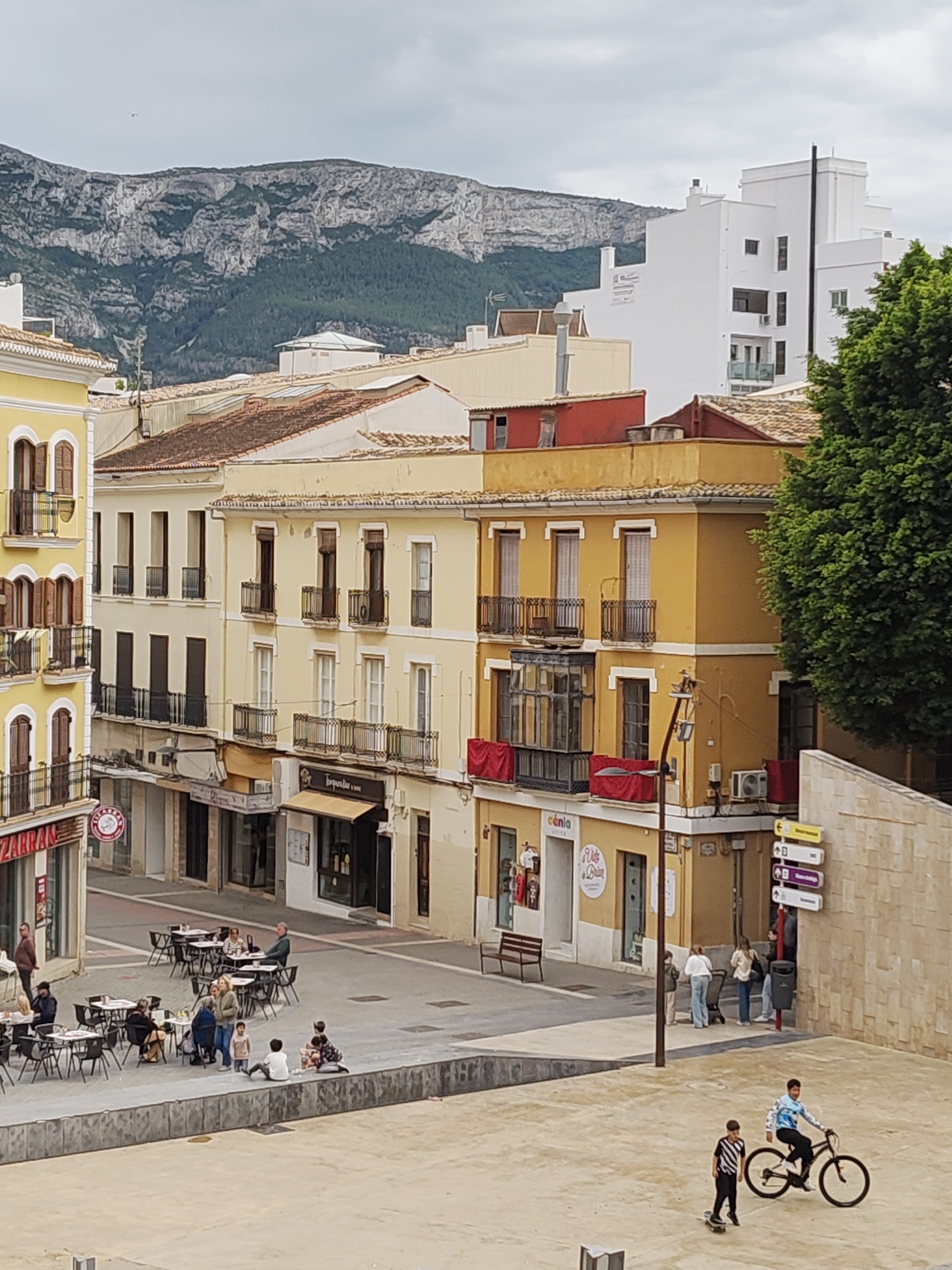 The image captures a cityscape in Dénia, Spain. The foreground features a public square with people engaging in various activities. Two boys are prominent; one is on a bicycle, and another rides a skateboard nearby. Other people are sitting at tables outside a restaurant and standing along the edge of the square.
In the background, the architecture consists of yellow and tan buildings with balconies and traditional Spanish features. A white building towers above the others to the right. In the distant background, a mountain range adds to the city's scenic environment. The sky is overcast. The image provides a glimpse into everyday life in this Spanish city.
