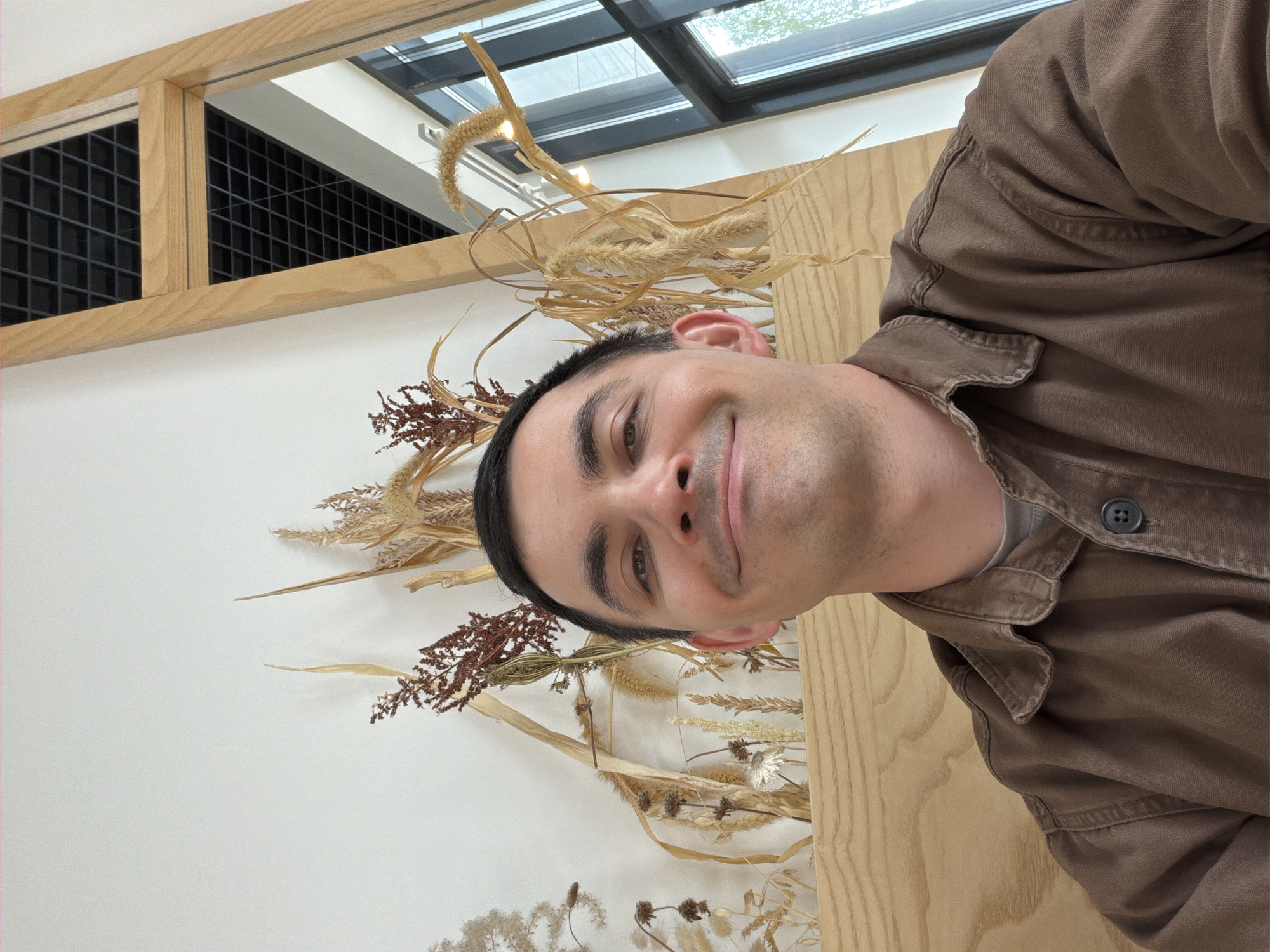 A man is taking a selfie. He is wearing a brown collared shirt. Behind him is a wall decorated with dried flowers and foliage. There is a window frame in the background with a view of what appears to be an indoor structure. The location is not discernible from the given image and therefore the city of Izhevsk, Russia cannot be confirmed.