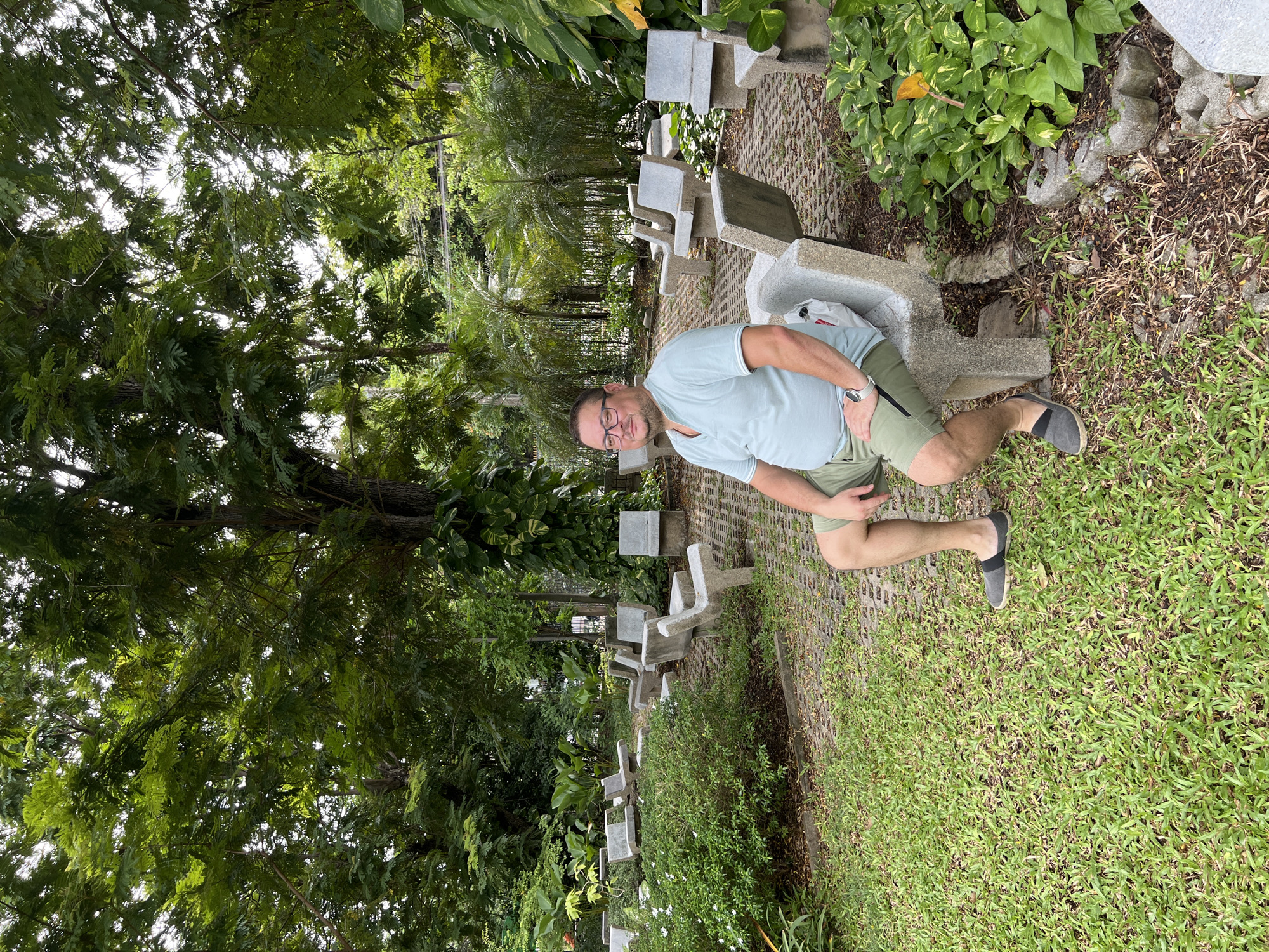 A man with glasses and a light blue polo shirt is sitting on a concrete bench in a park. He is wearing green shorts and dark gray slip-on shoes. In the background, there are more concrete benches arranged in rows, as well as lush green trees and foliage. The ground is a mix of grass and a paved walkway made of interlocking concrete blocks. The overall scene suggests a peaceful outdoor setting within the city. This scene is located in Bangkok, Thailand.
