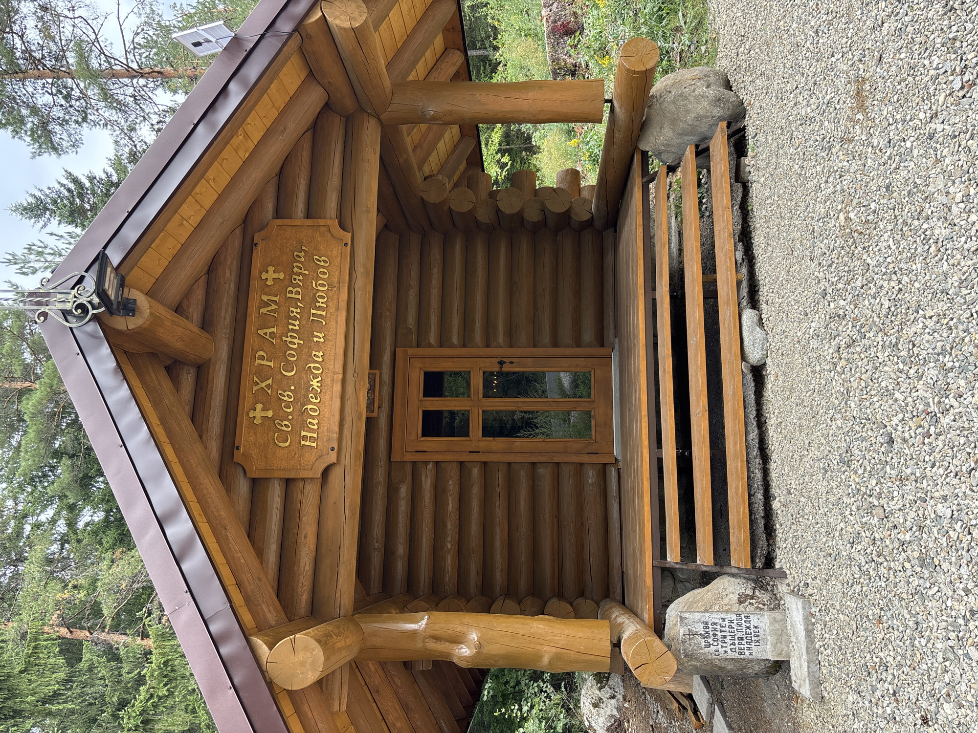 The image shows a small, rustic wooden church. The church is constructed from logs, with a brown metal roof. A sign above the entrance reads "ХРАМ СВ. С. СОФИЯ, ВЯРА, НАДЕЖДА И ЛЮБОВ" (Church of St. Sofia, Faith, Hope and Love). There is a wooden door with glass panes leading into the church, and a set of wooden steps leading up to the entrance. A stone marker with inscriptions is located to the left of the steps. The church is situated in a natural setting, surrounded by trees and gravel. In the distance, there is foliage and more trees. The lighting suggests it is daytime.