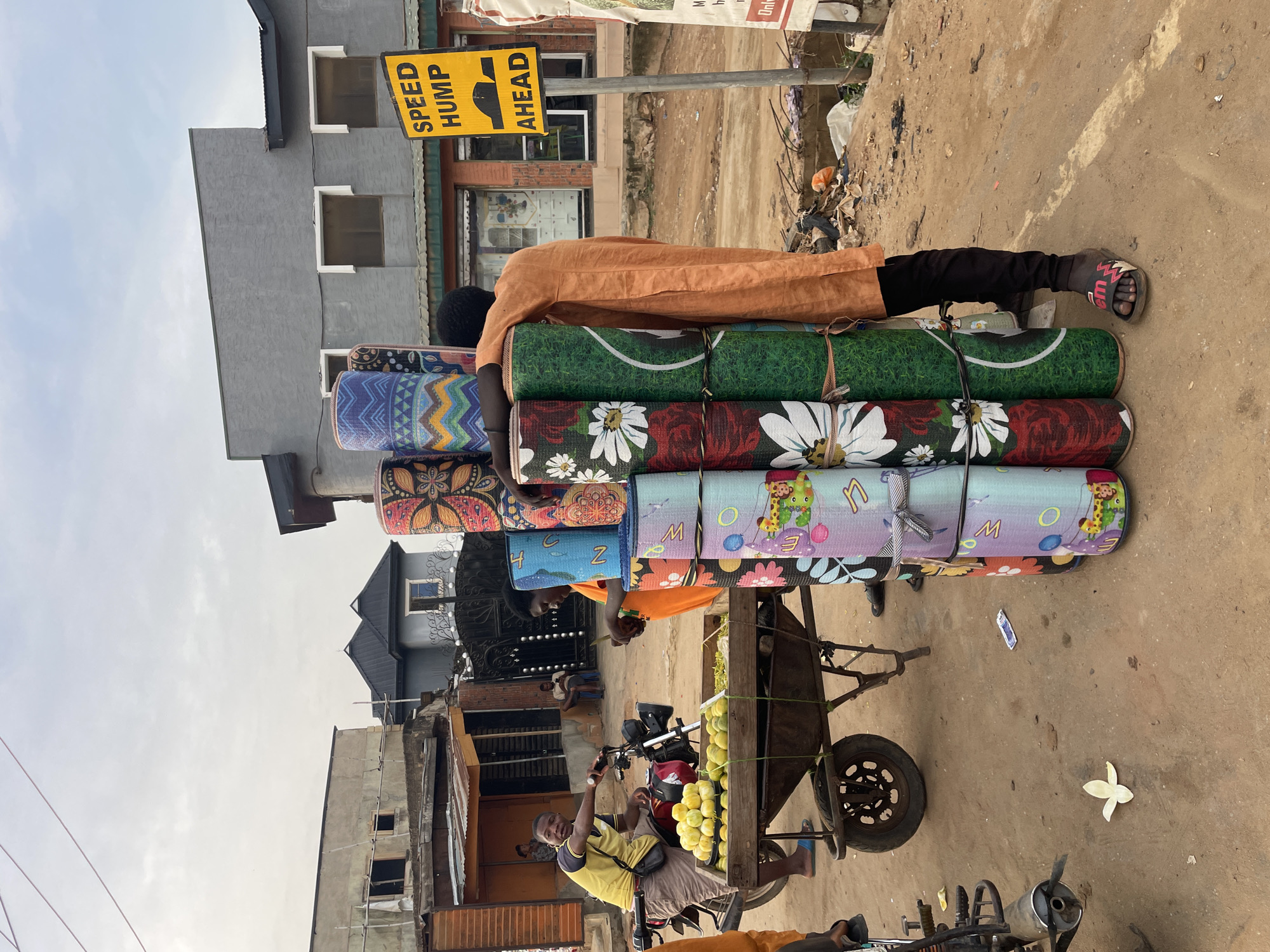 A street scene in the city of Unknown, Unknown, shows a man carrying a stack of rolled mats. The mats have various designs, including geometric patterns, floral motifs, and cartoon characters. The man is wearing an orange shirt and black pants. A "Speed Hump Ahead" sign is visible in the background. In the lower part of the frame, a vendor on a motorcycle is seated next to a cart loaded with melons. The environment appears to be an urban area with buildings and other street vendors.