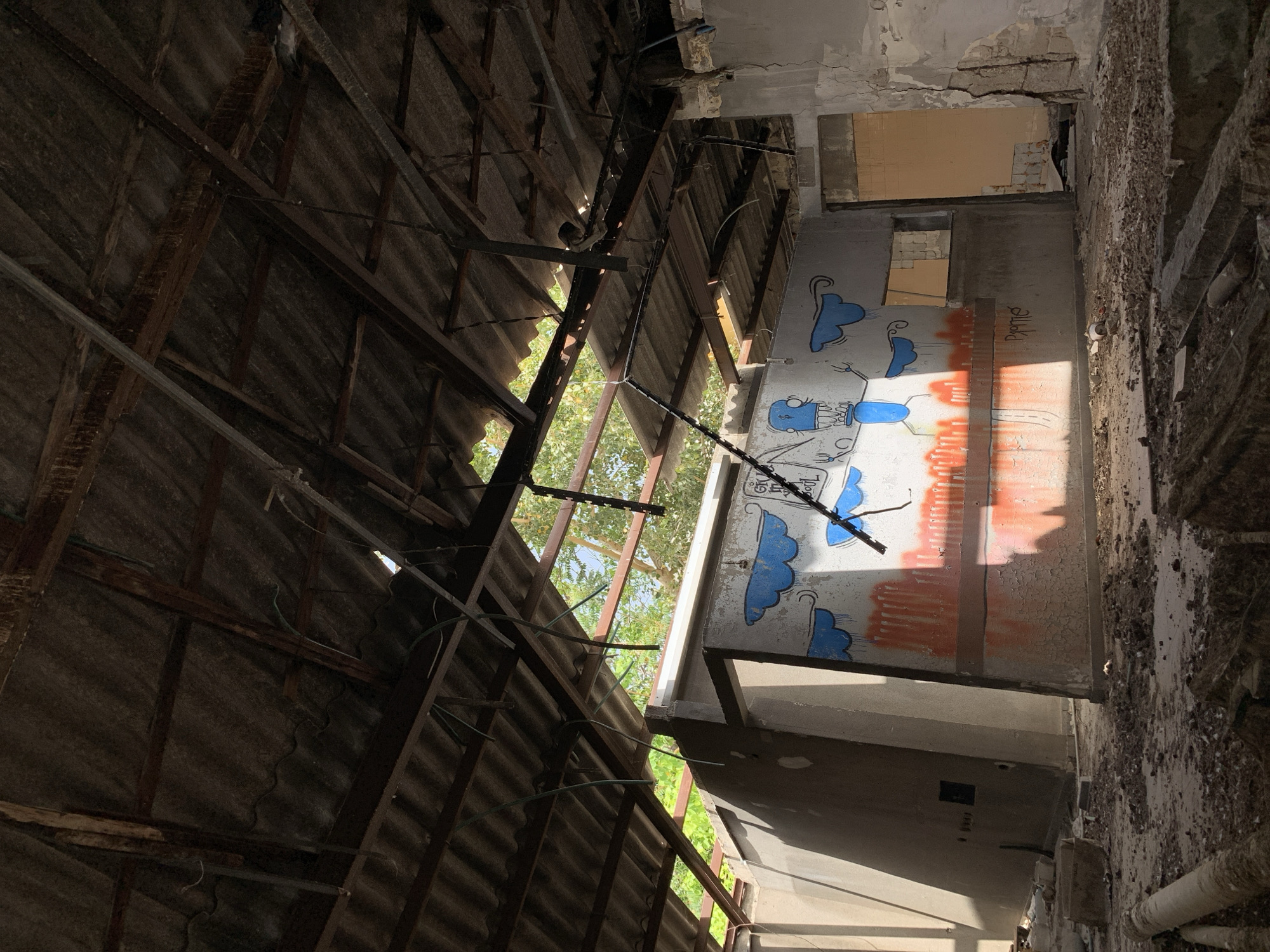The image depicts the interior of a dilapidated building in Tel-Aviv, Israel. The structure shows significant signs of disrepair, with a partially collapsed corrugated metal roof exposing the sky and foliage. The walls are decaying, covered in graffiti, including blue clouds and stylized figures. Debris litters the floor, and structural elements show signs of deterioration. The overall scene suggests abandonment and neglect.