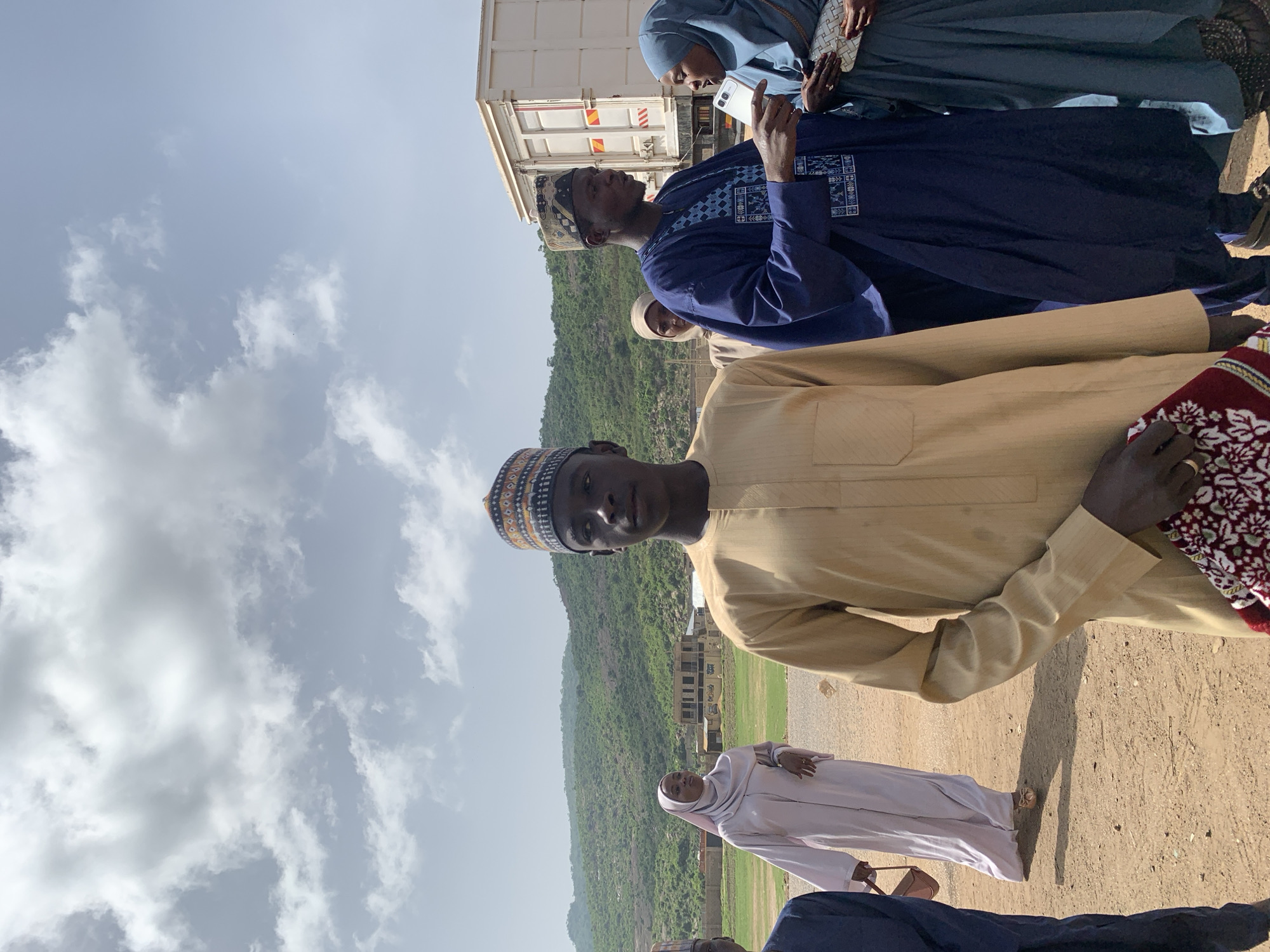 The image shows a group of people in what appears to be the outskirts of Naraguta Hausawa, Nigeria. The foreground features several individuals in traditional Hausa clothing, including a man in a tan robe and a patterned cap prominently facing the camera. To his right, partially visible, is someone in a dark blue garment with patterned embroidery. Further back, a woman in a blue hijab is holding a smartphone. A woman in a light-colored garment walks behind the group.
In the background, a truck is parked. Beyond the truck, the terrain slopes up to a hill covered in vegetation. An unfinished building can be seen on the hillside. The ground is dusty. The sky above is cloudy.