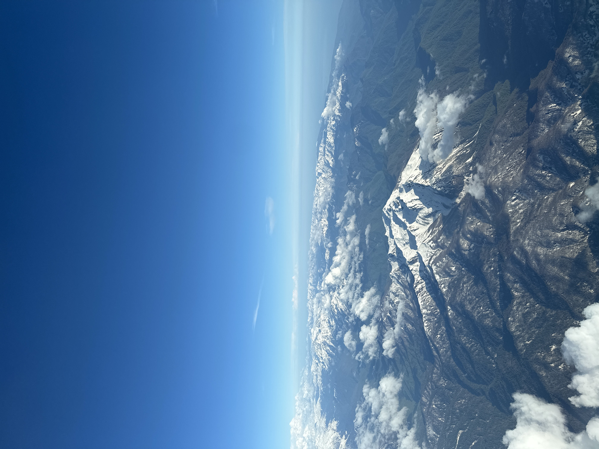 The image shows a landscape with mountains covered in snow and patches of forest, viewed from an aerial perspective. Clouds are interspersed among the mountain peaks. The sky is a clear blue, with a lighter horizon indicating distance or atmospheric haze. The image does not depict the city of Khimki, Russia.