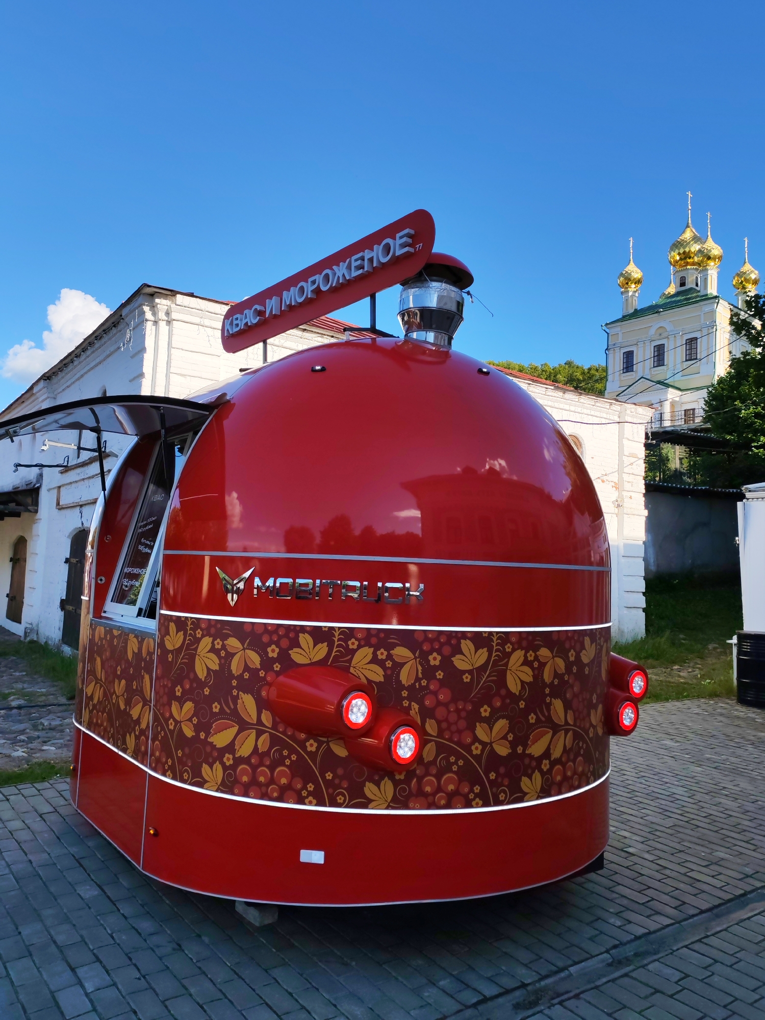 This media file shows a bright red, egg-shaped food truck parked on a cobblestone surface. The truck has a decorative pattern of golden leaves and red berries on its lower half. It features a prominent sign in white lettering that reads "КВАС И МОРОЖЕНОЕ" (Kvass and Ice Cream). On the side of the truck, partially visible through an open serving window, are handwritten signs listing prices in rubles for "КВАС" (Kvass) and "МОРОЖЕНОЕ" (Ice Cream). The word "МОБИТРУК" (MOBITRUCK) is displayed in metallic lettering on the side of the truck.
In the background, a traditional Russian church with golden domes stands against a clear blue sky. The church is light yellow with white architectural details. The scene appears to be set in Plyos, Russia, given the architectural style of the church and the presence of a traditional food truck. The time of day is likely midday, as indicated by the bright sunlight and clear sky. There are no people visible in the frame. The overall impression is one of a bright, sunny day in a picturesque, historic setting, with a modern food service element added.