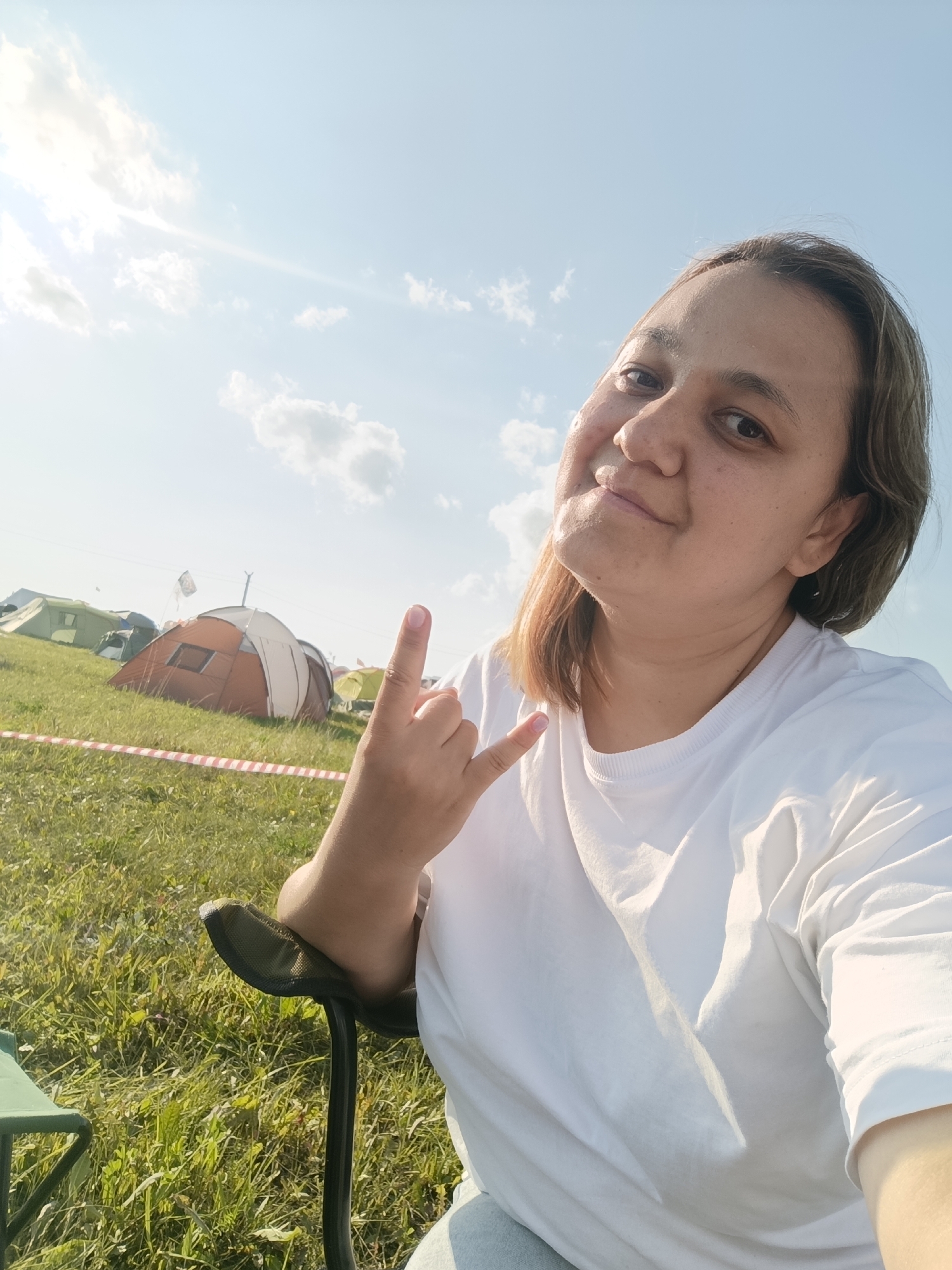 A woman with medium-length brown hair and a light complexion is sitting in a black outdoor chair, holding her right hand up in a rock-and-roll gesture. She is wearing a white t-shirt. The scene appears to be a campground with several tents visible in the background, situated on a grassy hill under a bright blue sky with scattered clouds. A red and white striped caution tape is visible in the foreground, bordering a section of the grass. The location is identified as Lagunovo, Russia.