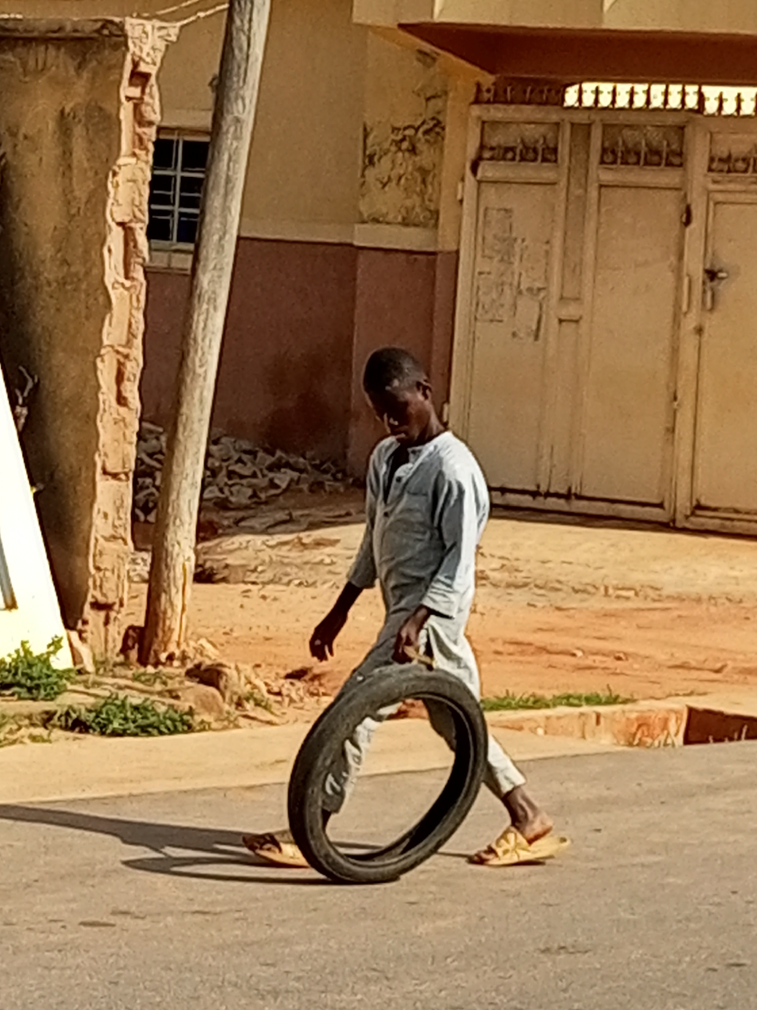 A young person, likely male, is depicted in mid-action in the city of Gombe, Nigeria. He is wearing a light grey, loose-fitting garment and yellow open-toed sandals. The individual is pushing a large, black tire along a paved road. His body is tilted forward, and his right leg appears to be extended to propel the tire. The setting includes a building with a damaged, textured wall on the left, revealing brickwork and a window with a grid. To the right of the building, there are large, light-colored double doors. The ground is a mix of paved road, reddish-brown dirt, and some sparse green vegetation. The overall scene suggests an outdoor, possibly urban environment.