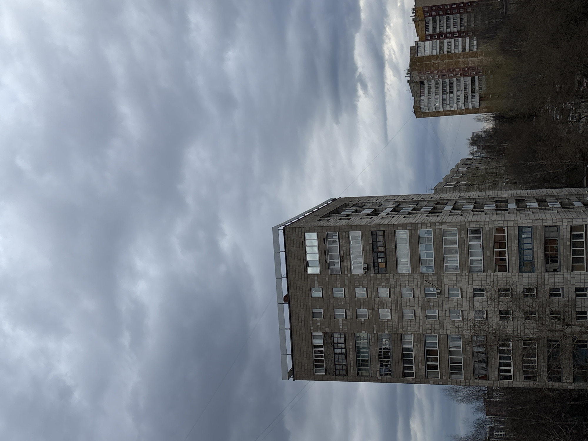 The image, taken with a vertical orientation and presented rotated 90 degrees clockwise, captures an urban scene featuring multi-story residential buildings under a heavily overcast sky. The weather is distinctly grey and cloudy, with thick, undulating cloud formations filling the entire sky, creating diffuse light. The time of day appears to be daytime, but without direct sunlight.
In the foreground, on the right side of the frame, stands a tall, grey apartment building constructed of what appears to be concrete blocks or large bricks. Its facade is punctuated by numerous windows of various sizes and several balconies, some enclosed. An external air conditioning unit is visible on one of the upper floors. A thin wire or power line stretches diagonally across the sky from the vicinity of this building.
Behind and to the left of the prominent foreground building, another multi-story residential block is partially visible through a cluster of bare trees. This building has a lighter, somewhat yellowish-brown or ochre-colored facade with more prominent, white-framed balconies. The trees surrounding the buildings are leafless, suggesting either late autumn, winter, or early spring. Hints of further urban structures are visible in the distant background.
The scene conveys a muted and subdued atmosphere due to the grey skies and bare trees. No people or specific activities are visible in the static cityscape. There is no discernible text present in the image. The architectural style of the residential blocks is characteristic of urban environments in Russia, aligning with the given location context of Perm.