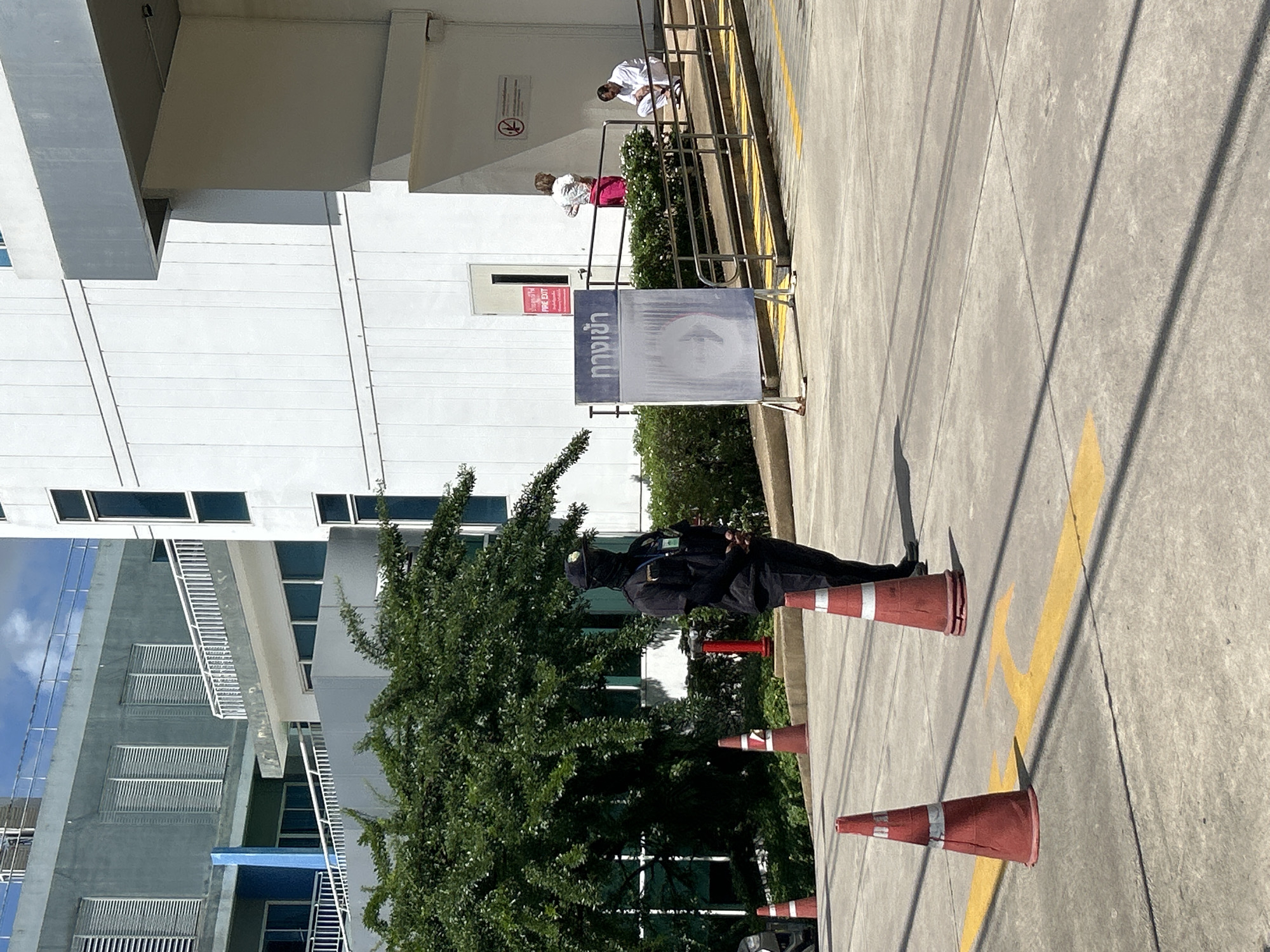 The image shows a street scene in Wichit, Thailand, with a security guard standing in the foreground, wearing a dark uniform and a face mask. The guard is positioned near several red and white traffic cones. Behind the guard, a large green tree is visible, as well as the exterior of a modern white building. To the right of the building, a ramp leads up to an entrance, with two people on the ramp and one person sitting near the top. A sign with Thai writing and "FIRE EXIT" written in English is posted on the building. The ground is paved, and yellow arrows are painted on the surface, indicating traffic direction. The sky is blue with some clouds.