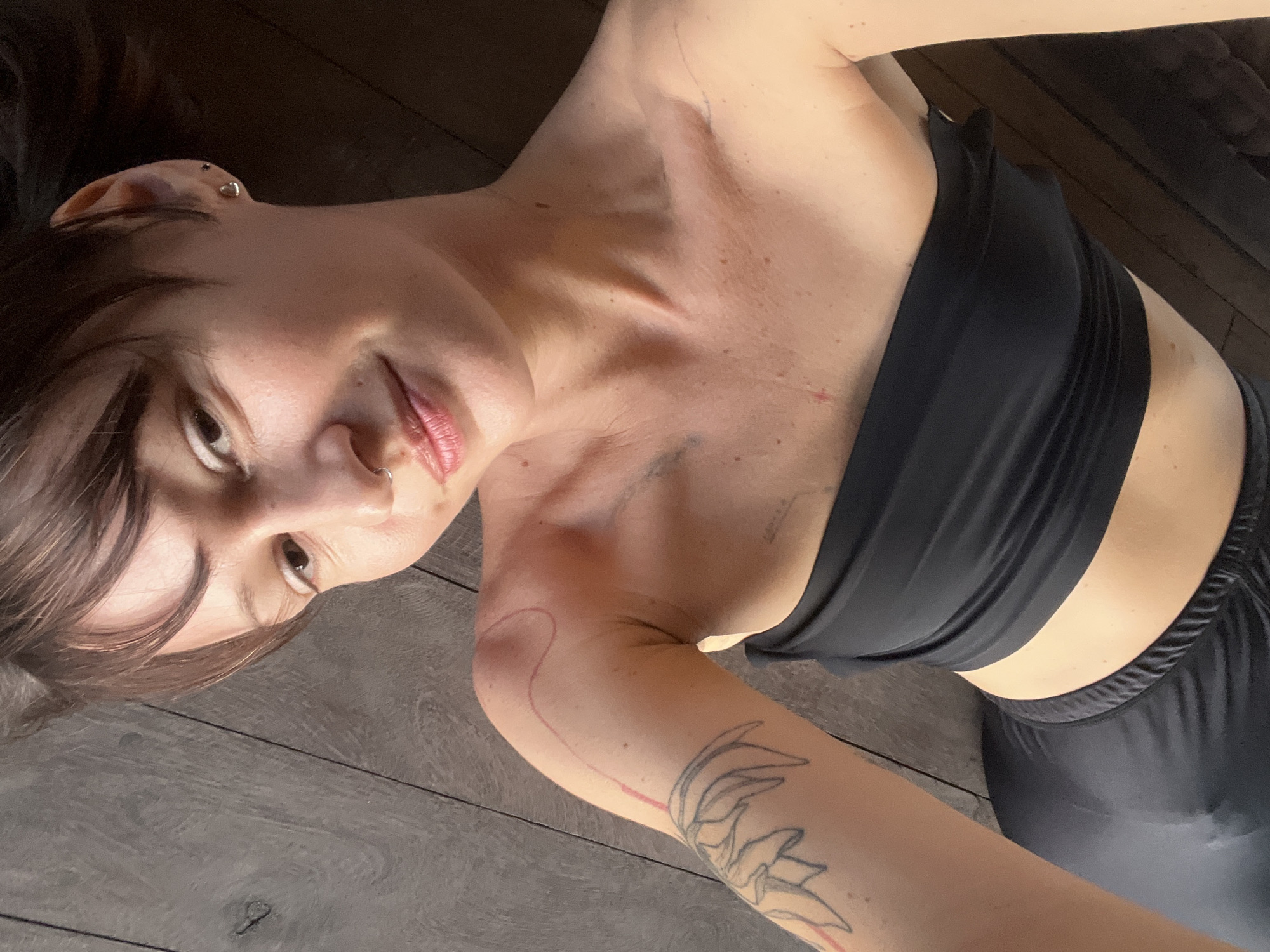 An overhead, medium shot features a person lying on their back on a dark wooden floor, looking up and slightly to the right with a soft smile. The person has shoulder-length dark brown hair, fair skin, and appears to be wearing a black strapless top or bralette and possibly matching black leggings or shorts.
Notable details include:
* **Facial features:** A nose ring (hoop) on the left nostril and what appears to be a small scar or mark above the right eyebrow. The lips are lightly colored.
* **Ear piercings:** A heart-shaped stud and another small stud are visible on the right earlobe and helix respectively.
* **Body details:** Several faint red marks or scratches are visible on the chest and neck area. A subtle line drawing tattoo, possibly a floral or leaf design, is visible on the upper left arm, extending down towards the forearm, with a curved red line drawn on the shoulder.
* **Clothing:** The black top has a ruched or ribbed texture.
* **Setting:** The dark wooden floor is the primary background, suggesting an indoor setting. The lighting appears natural, indicating daylight.
The overall mood is relaxed and candid. No visible text is discernable in the image. The location context is Mengwi, Indonesia.