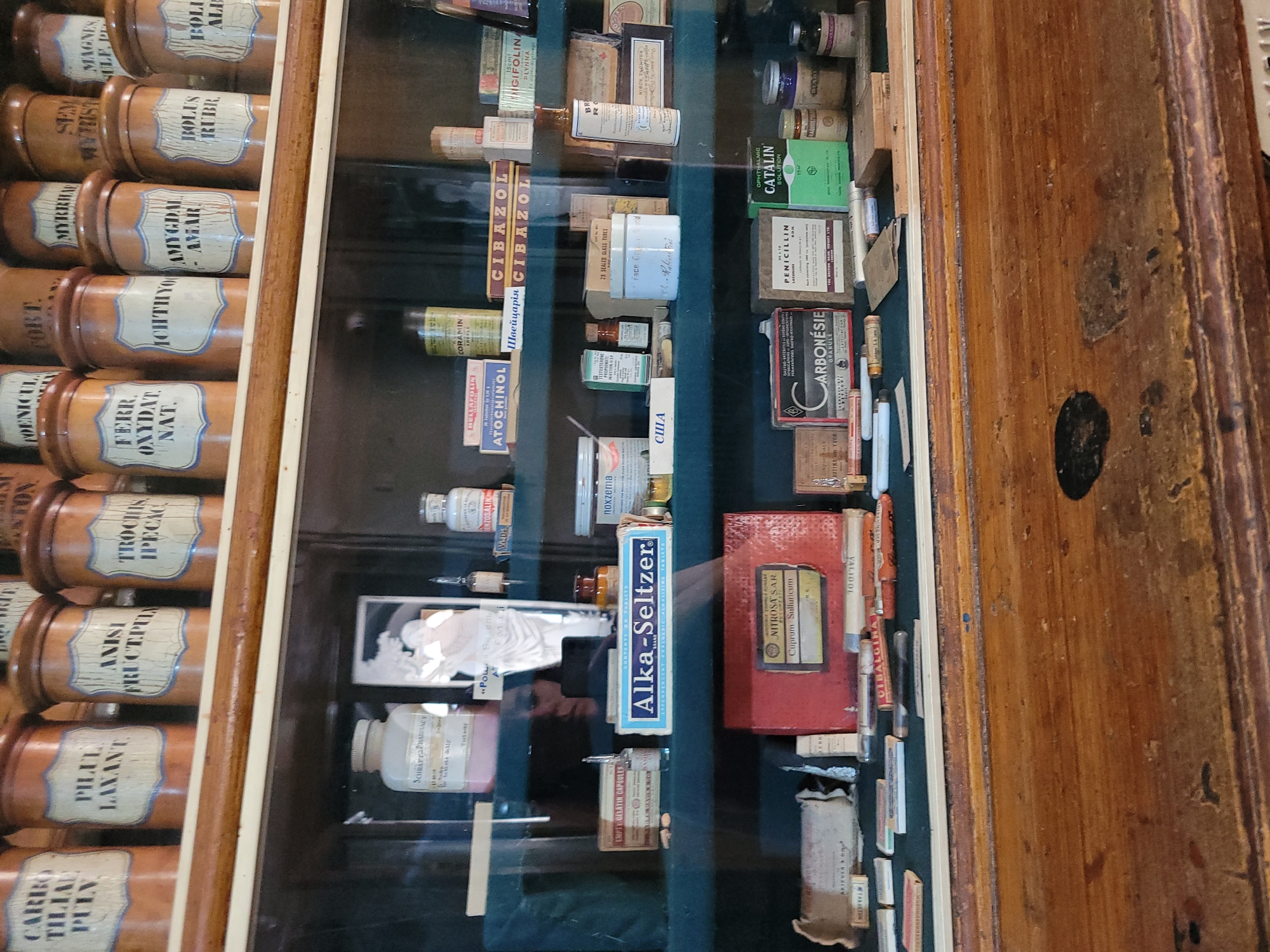 The image displays an antique apothecary cabinet filled with various vintage medicinal products. The top shelf contains rows of brown ceramic jars with handwritten labels, such as "CARRO THILAU PULV.", "PILUL LAXANT.", "ANISI FRUCT. PULV.", "TROCHIS IPECAC.", "FERR. OXYDAT. NAT.", "ICHTHYOL", "AMYGDAL AMAR.", "BOLUS RUBR.", and "BOLUS ALIN.". Below, the second shelf holds glass bottles, boxes, and vials. Notable items include a pink-labeled bottle, a framed photograph, an Alka-Seltzer box, and several smaller boxes and bottles. The third shelf features more packaged medicines and vials, including a red box labeled "NITROSA'S AR. Cuprum Sulfidicum", boxes of "CARBONÉSIE GRANULE" and "PENICILLIN LOZENGES", and a green box of "CATALIN SOLUTION". The items are arranged on dark shelves within a wooden cabinet, illuminated by ambient lighting. The environment appears to be an indoor setting, likely a museum or a preserved historical space.