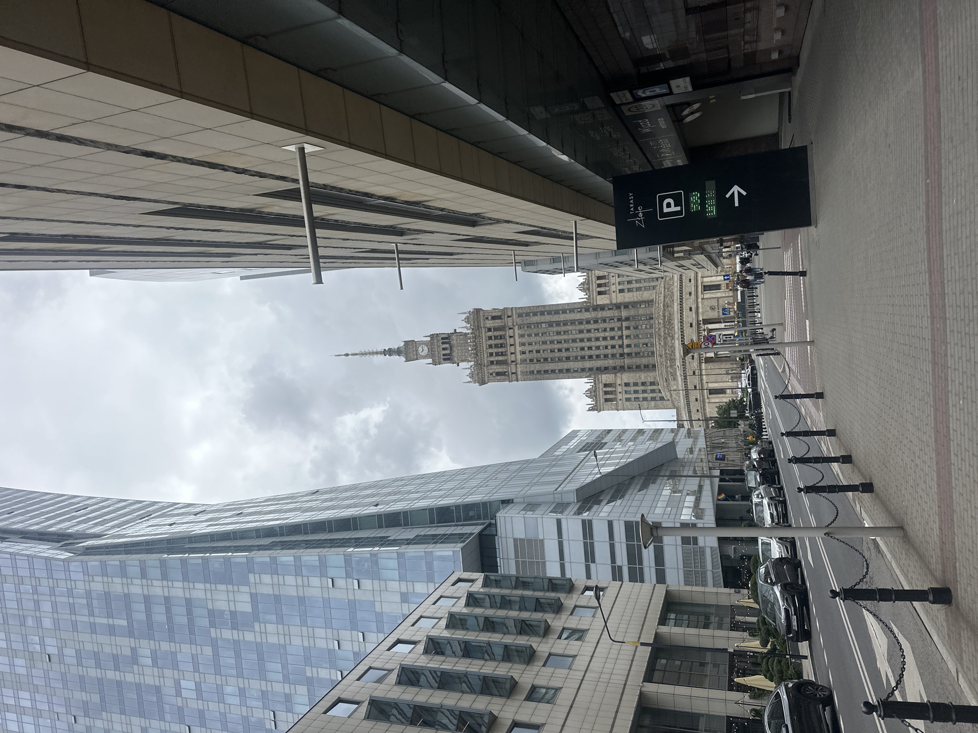 This image shows a street scene in Warsaw, Poland, looking towards the Palace of Culture and Science. On the left, a modern glass skyscraper with a curved facade dominates the view. Adjacent to it is a beige building with a grid of windows. The street itself is paved with gray and red bricks, with a black metal chain-link fence separating the sidewalk from the road. Several cars are parked along the street. On the right, a contemporary building with a beige tiled facade features a dark, angled sign that reads "TARASY ZIELONE" and indicates parking. In the background, the iconic Palace of Culture and Science, a towering Stalinist-era skyscraper, stands tall against a cloudy sky. Several people are walking on the sidewalk and crossing the street in the distance. The overall atmosphere is urban and overcast.