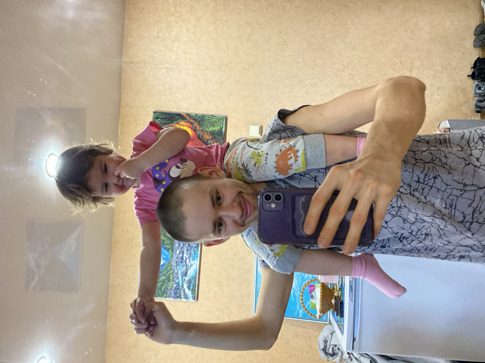 A man is giving a child a piggyback ride and taking a selfie with a purple phone. The child is a toddler wearing a pink t-shirt. The man has short hair and is wearing a grey patterned shirt. The background shows a tan wall with three framed pictures. A white refrigerator is partially visible. There is no confirmed information that the location is in the city of Bely Klyuch, Russia.