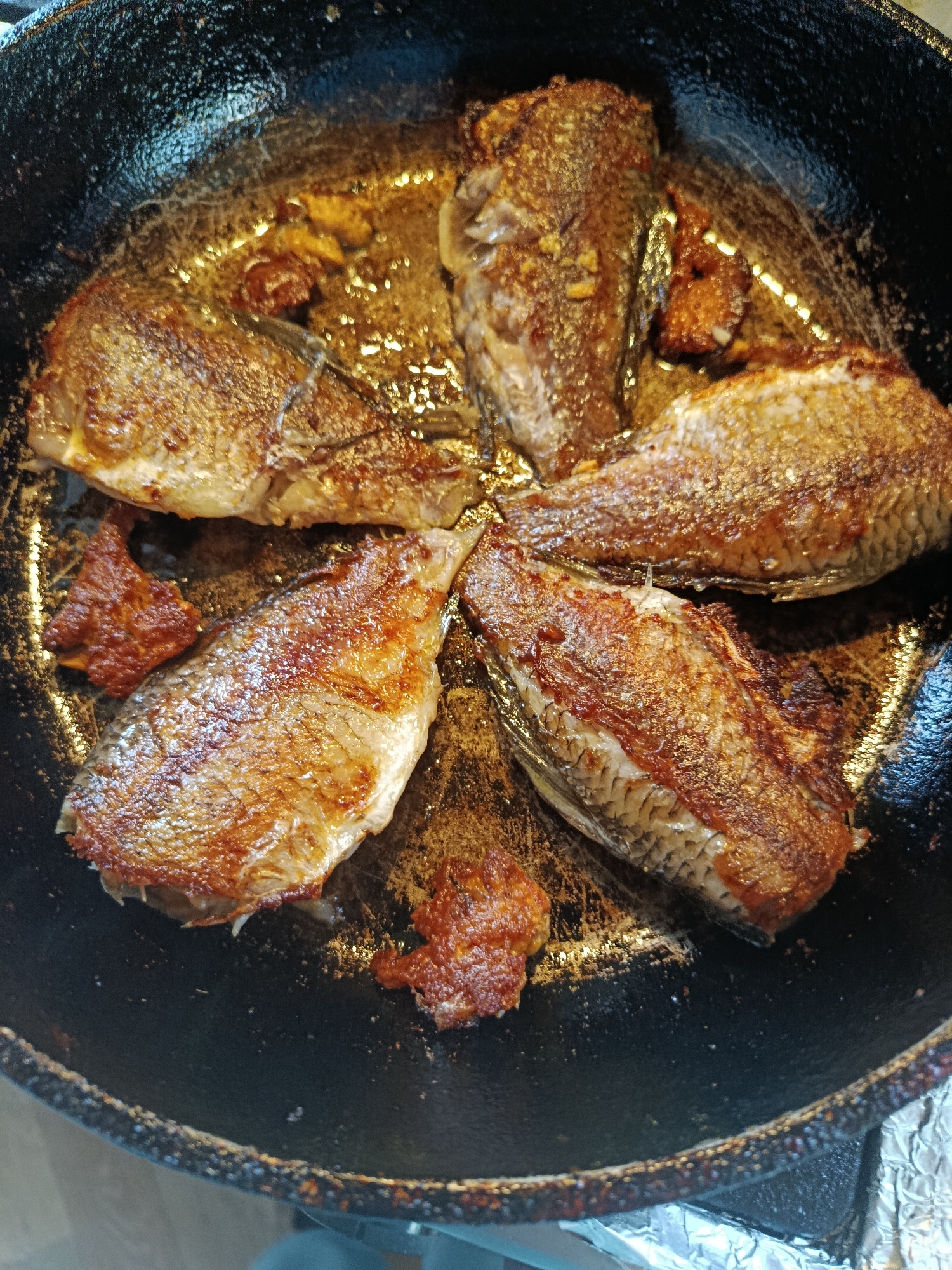 In a dark, cast-iron frying pan, five whole, small, whole fish are arranged in a circular pattern. The fish are golden brown and appear to be pan-fried, with visible scales and some crispy bits. Small pieces of fried food, possibly the same fish or another ingredient, are scattered around the fish. The pan is filled with glistening oil, reflecting the light. The background is dark and out of focus. The scene is not indicative of a specific city or location.