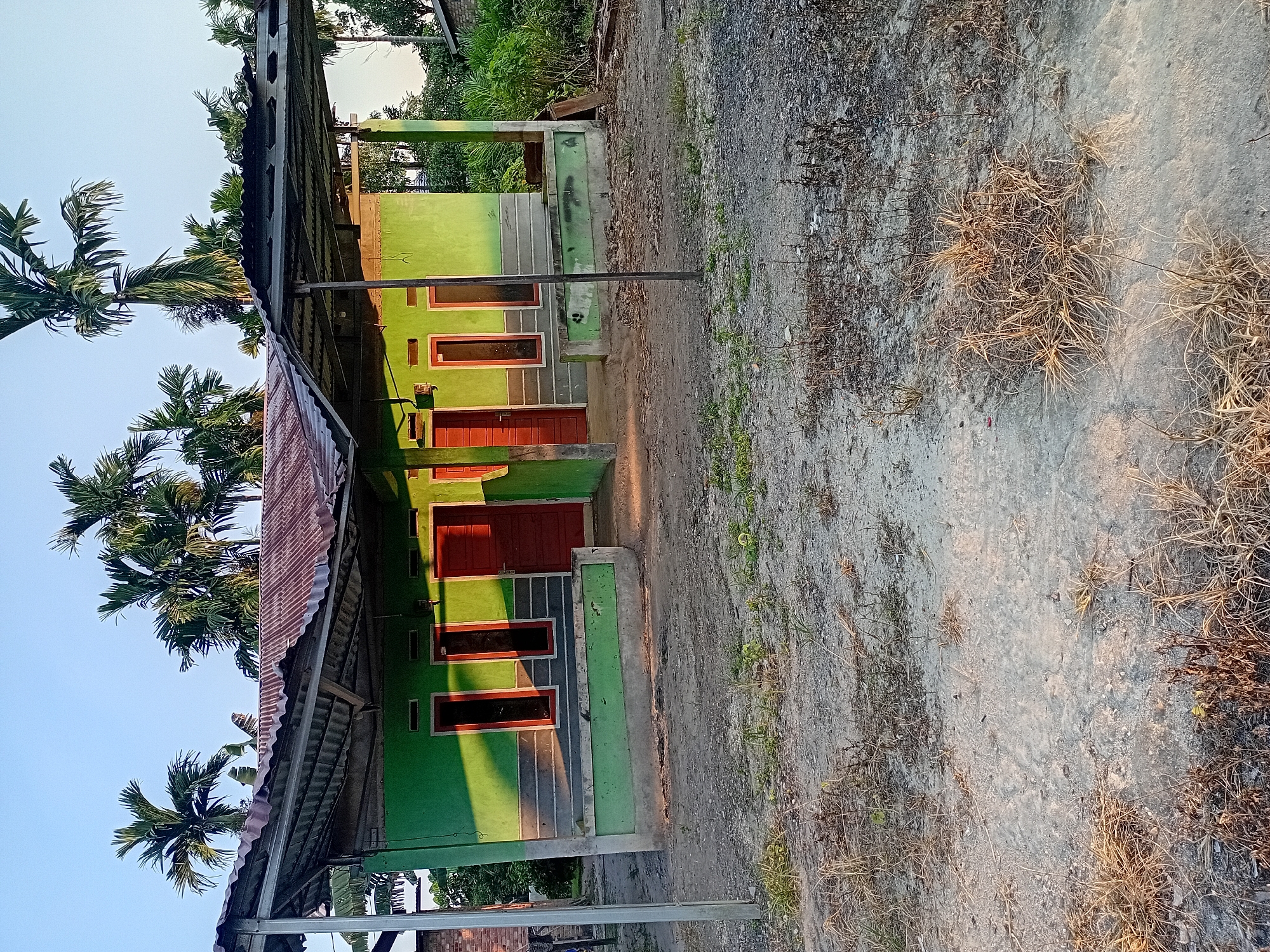 The image shows a single-story, bright green house with a corrugated metal roof, characteristic of construction in Kasang Pudak, Indonesia. The house has several prominent red doors and windows with red frames. A covered porch area extends from the front of the house, supported by metal poles. The ground in front of the house is dirt and gravel, with patches of dry grass. In the background, tall palm trees are visible, suggesting a tropical or subtropical environment typical of the region. The sky is clear and blue, indicating it is daytime. The scene appears to be a residential area.