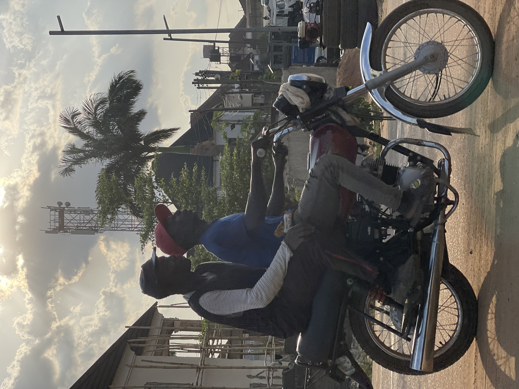 Two individuals are situated on a red motorcycle, parked on a paved surface. The person on the front of the motorcycle is wearing a blue shirt and a red cap. The person on the back of the motorcycle is wearing a black vest with white stripes, and a black cap. The location appears to be within a city, identifiable by various structures including a telecommunications tower, power lines, and buildings under construction. The scene seems to be in Abuja, Nigeria, but further validation is needed for the precise city location. The photograph is taken during daylight hours.