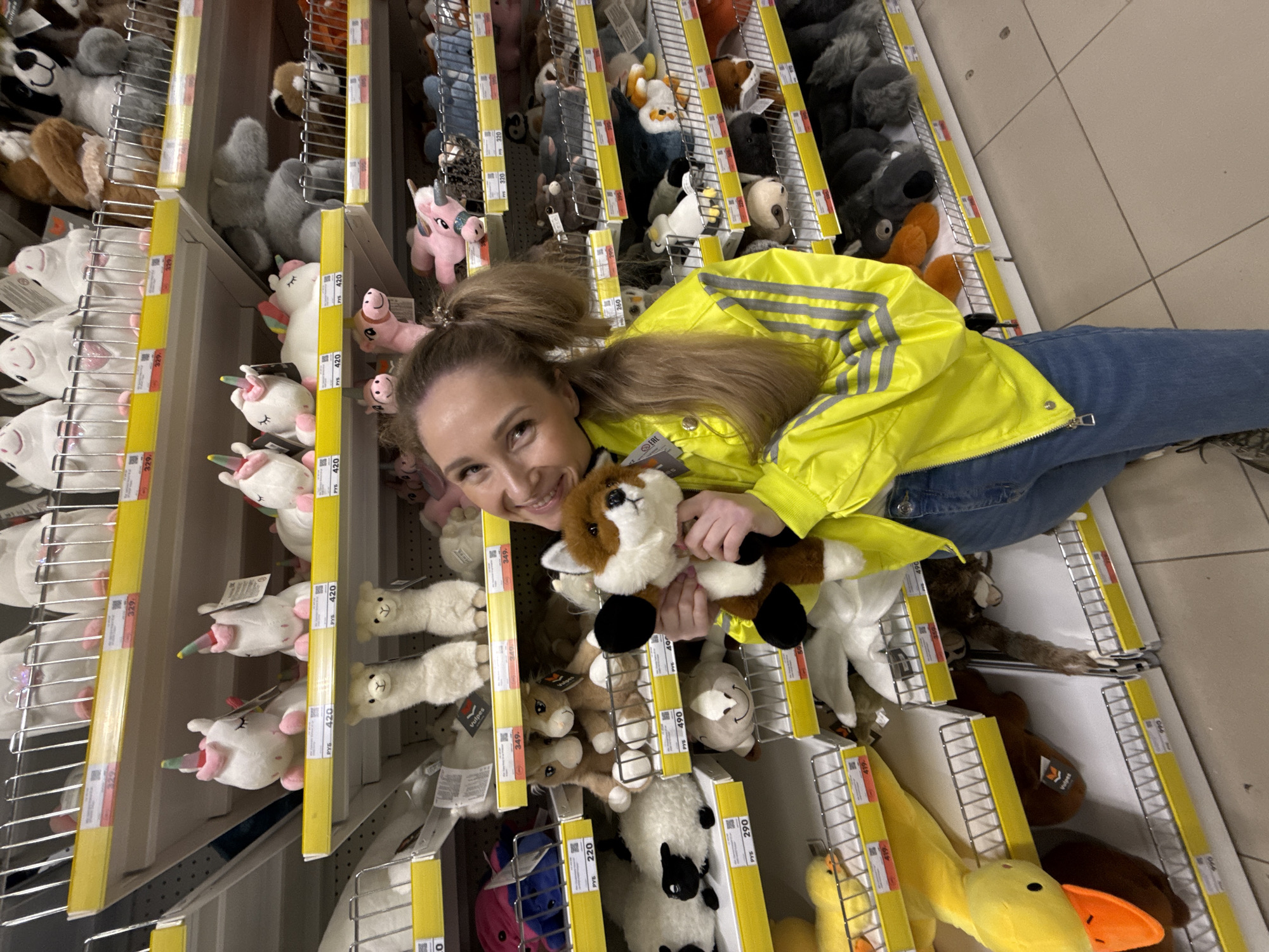 The scene is an indoor retail aisle in Izhevsk, Russia, displaying a wide variety of plush toys on white shelving units with yellow price strips. A female individual is positioned in the central-right of the frame, facing and smiling directly at the camera. She has light brown hair styled in two pigtails and wears a bright yellow zippered jacket with grey reflective stripes on the sleeves and shoulders, over blue denim jeans. She holds a brown and white plush fox toy with both hands. The shelves are densely stocked with diverse stuffed animals, including white unicorns, llamas, sheep, ducks, and foxes. Prices are visible in Russian Rubles (РУБ) on the yellow strips. The floor is a light-colored tiled surface.
