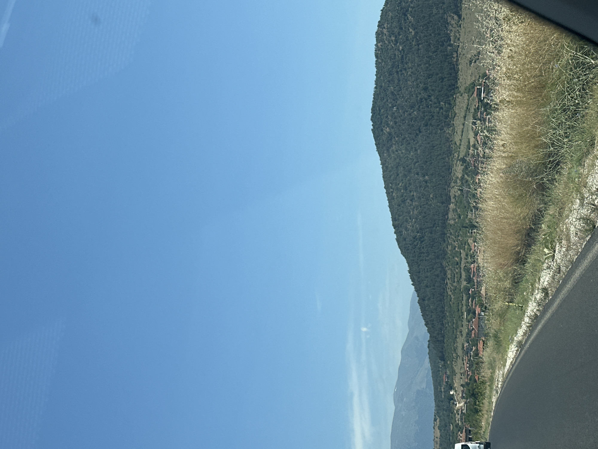 The image depicts a landscape scene with a paved road in the foreground curving to the left. On the right side of the road, dry, tall grass grows, interspersed with some greenery. Beyond the grass, a village with red-tiled roofs is visible nestled in a valley. Behind the village, a large, forested hill dominates the middle ground, with its slopes covered in dark green trees. Further in the background, softer, lighter green hills recede towards the horizon under a clear blue sky with a few wispy white clouds. A white van is partially visible on the left side of the road. The overall setting appears to be rural, likely in or near the city of Pesnopoy, Bulgaria, given the architectural style of the village and the mountainous terrain.