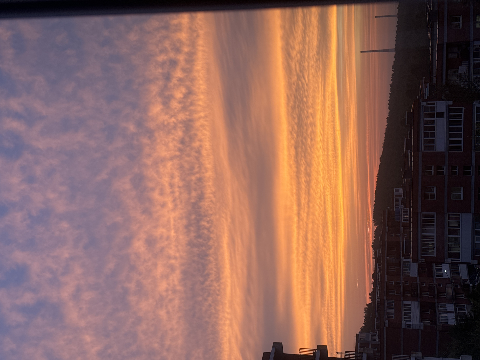 A panoramic view captures a vibrant sunset over Markova, Russia. The sky is a canvas of warm hues, with bands of orange and pink clouds stretching horizontally across the horizon, transitioning to a soft lavender and blue at the top. Below, the silhouettes of apartment buildings with red brick facades and white window frames form a dense urban landscape. These buildings are arranged in a clustered pattern, suggesting a residential area. A dark, tree-covered hill rises in the background, partially obscuring distant industrial structures, including two tall, thin smokestacks or chimneys. The overall scene conveys a sense of evening tranquility in an urban setting, illuminated by the dramatic natural light of the setting sun.
